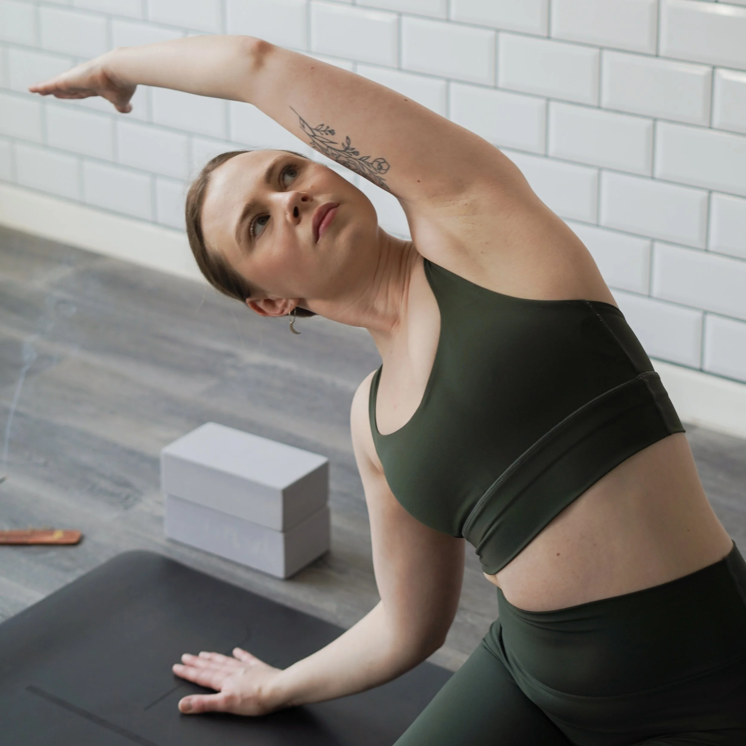 A woman doing a yoga pose on a black mat in a room with white tiled wall, wearing a black sports bra and green workout shorts.