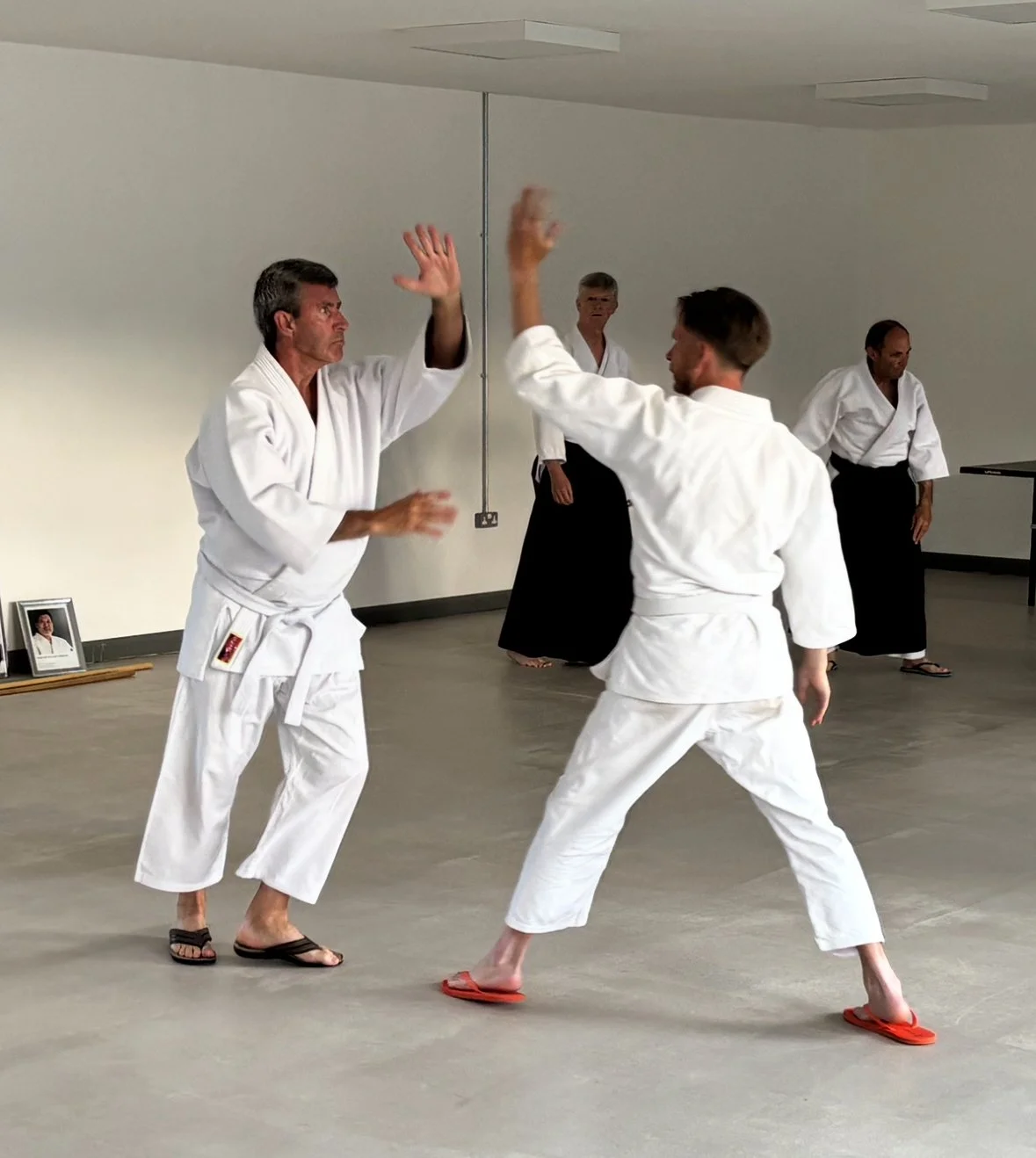 Two men practicing martial arts, wearing white traditional uniforms, sparring in a dojo with three other practitioners observing in the background.