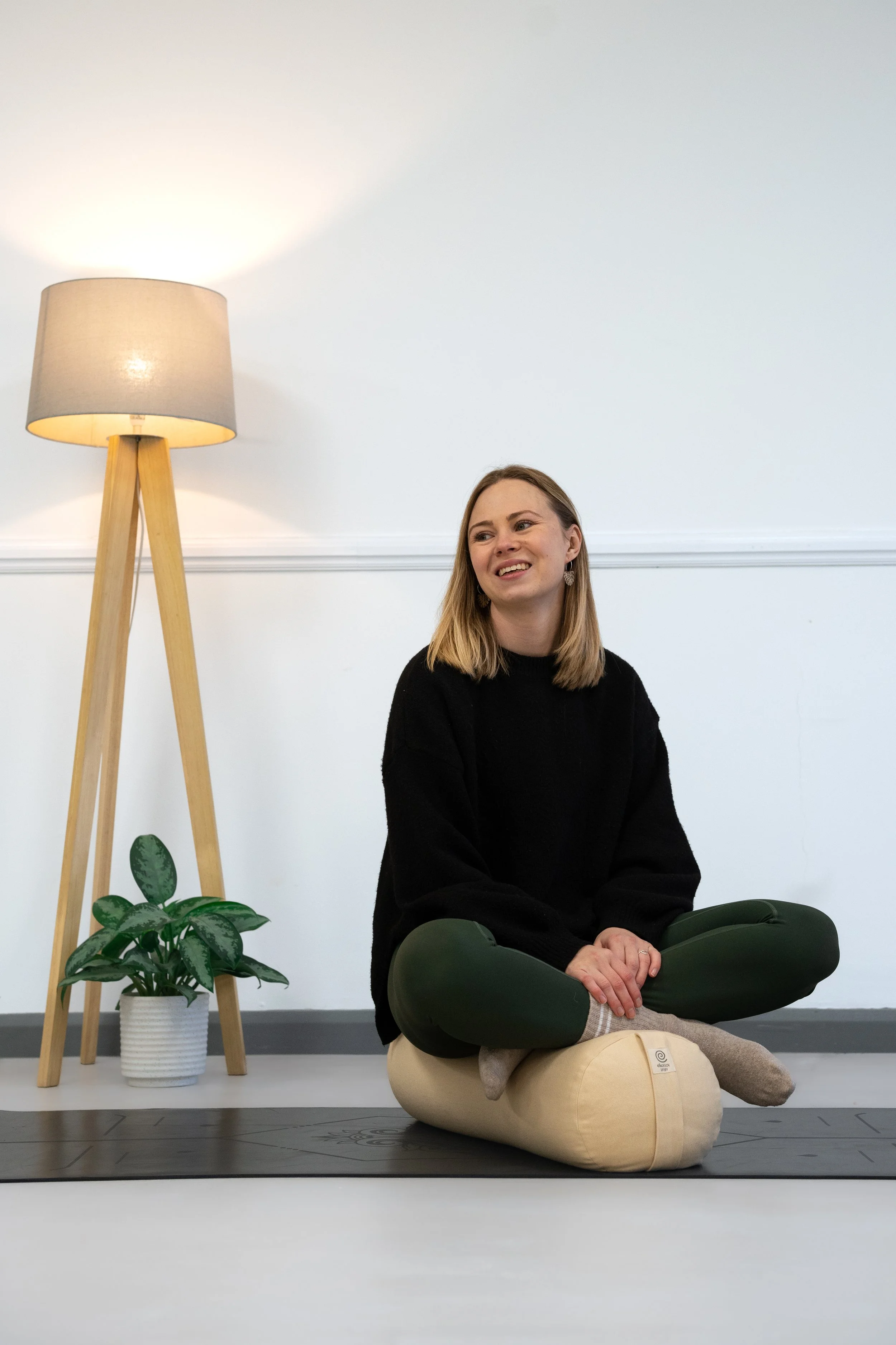 A woman with blonde hair sitting cross-legged on a yoga mat indoors, smiling while wearing a black sweater and green pants, with a lamp and a potted plant behind her.