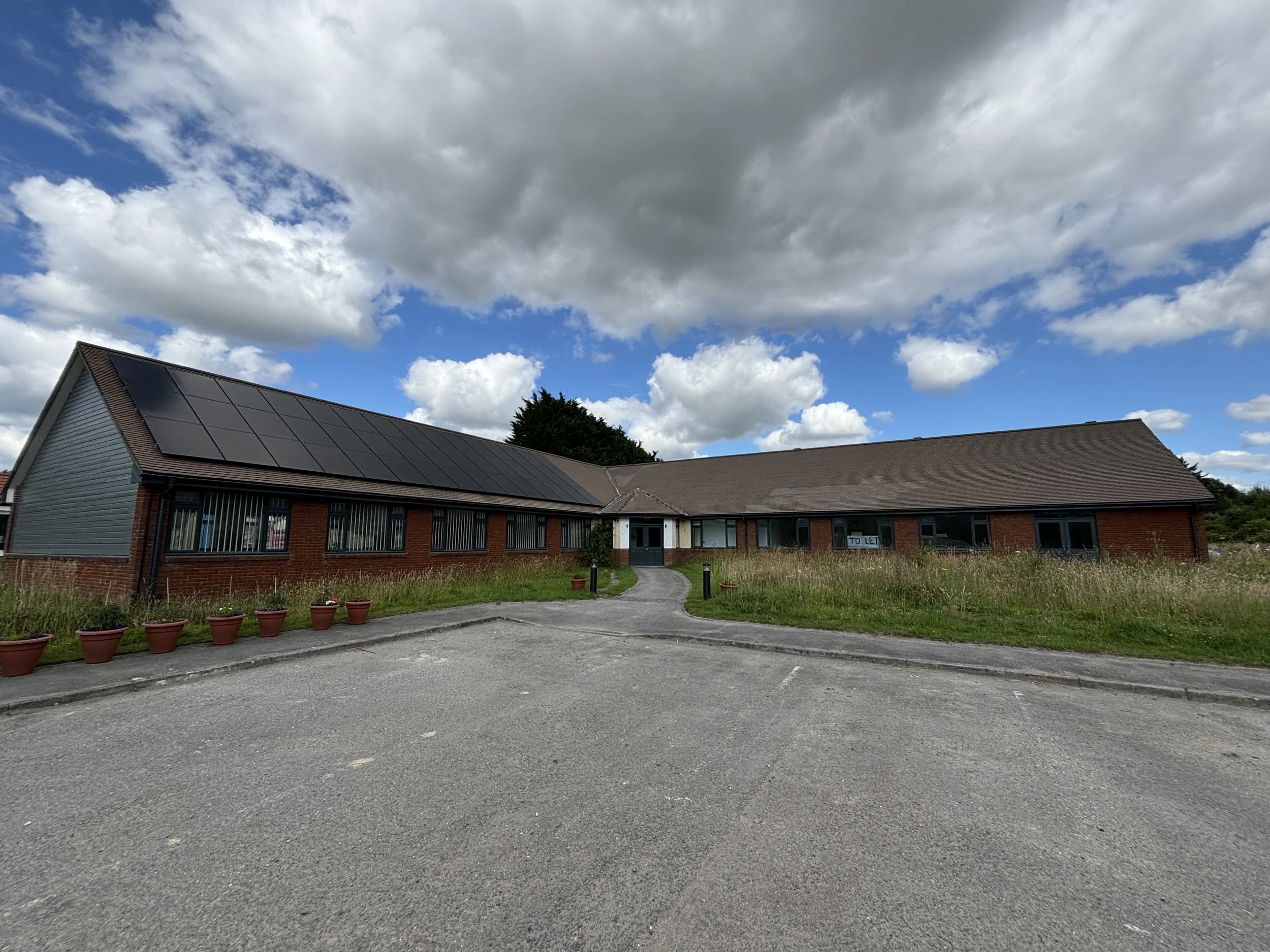 A parking lot in front of a building with solar panels on the roof. The sky above has large, partly cloudy clouds.