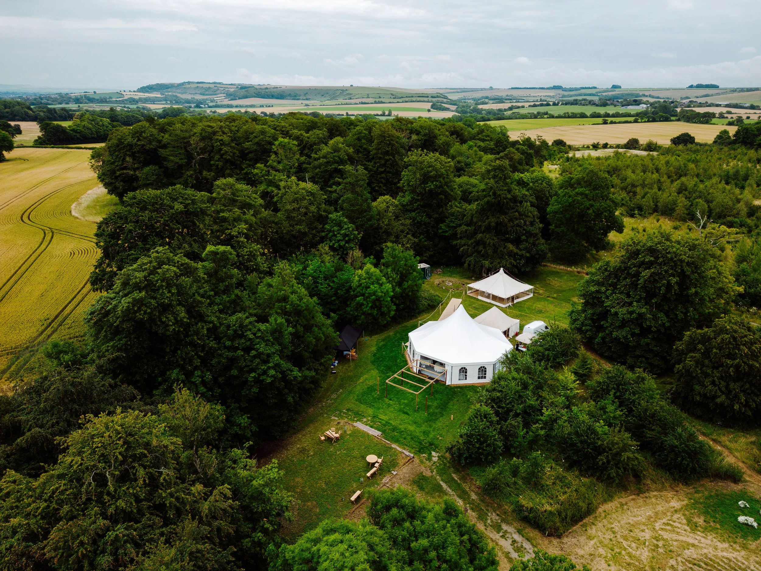 Aerial view of a countryside with large green trees and a white event tent with smaller structures nearby, surrounded by open fields and rolling hills.
