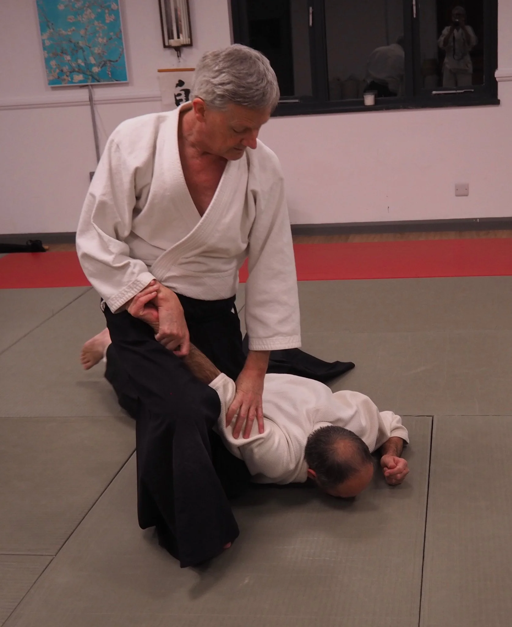 Two martial artists practicing a Judo or Brazilian Jiu-Jitsu technique on a blue martial arts mat in a dojo or training room, with one person in a white gi and the other in a white gi, in front of a wall with artwork and windows.