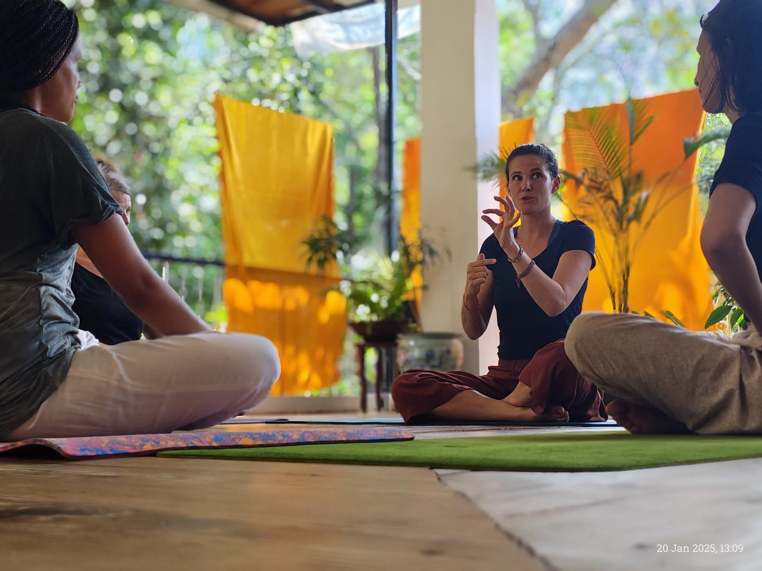 Group of people sitting on the floor in a circle, engaged in a discussion or meditation session indoors with large windows and greenery outside.