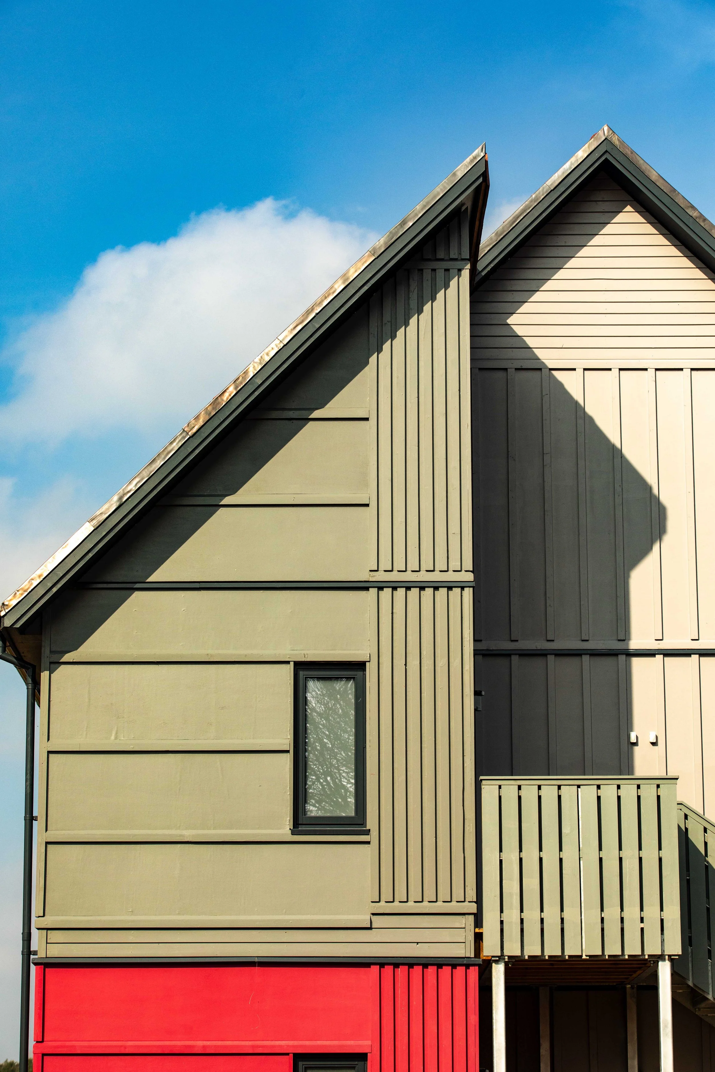 Close-up of a modern house exterior with beige wooden siding, a small window, and a red section at the bottom, under a blue sky with a few clouds.