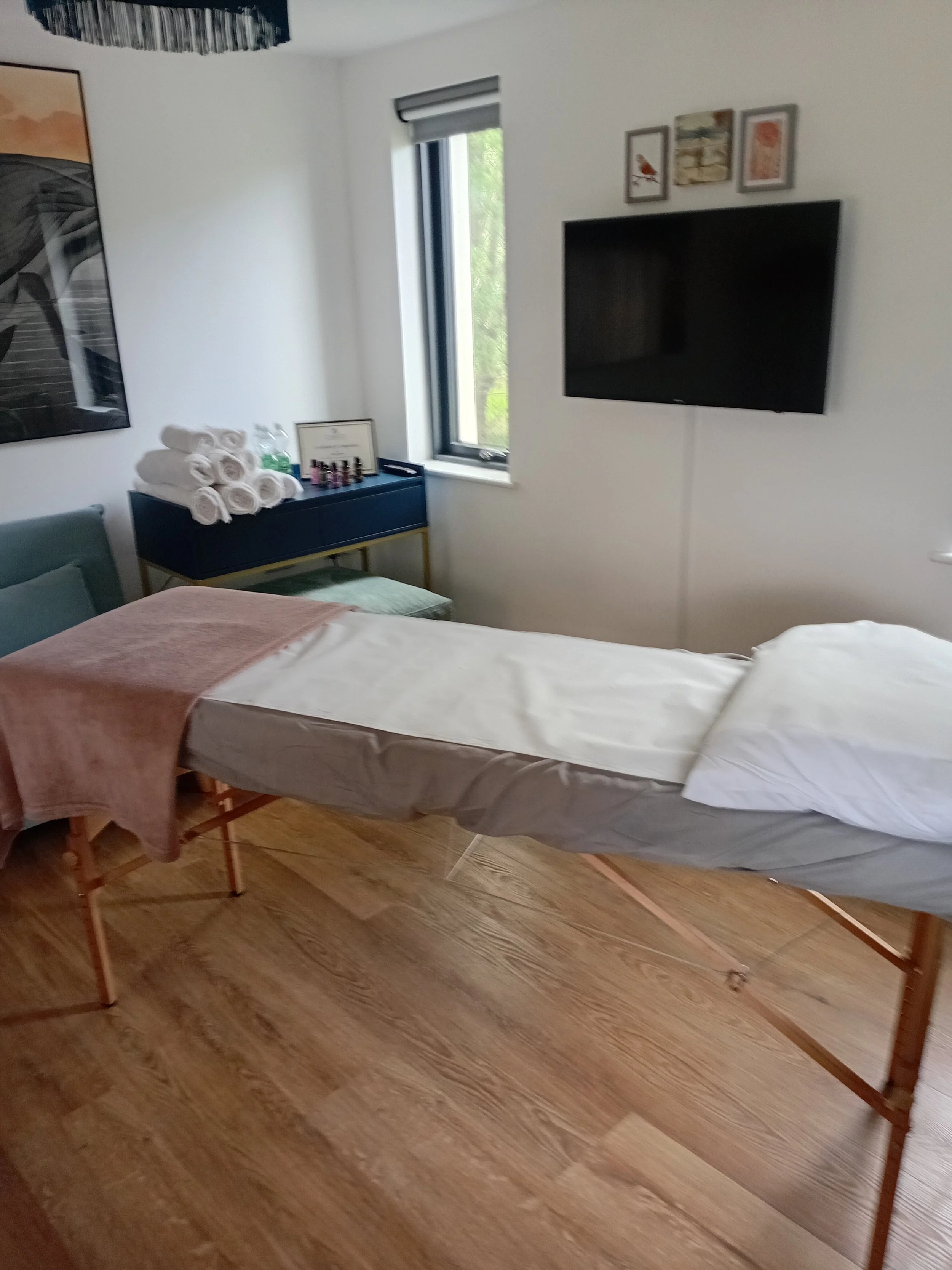 A massage table with white sheets and a pink towel. On a black side table are rolled white towels, two green bottles, a framed certificate, and a collection of nail polish bottles. A television is mounted on the wall, and three framed pictures are above it. A window with a partially open blind shows green trees outside.