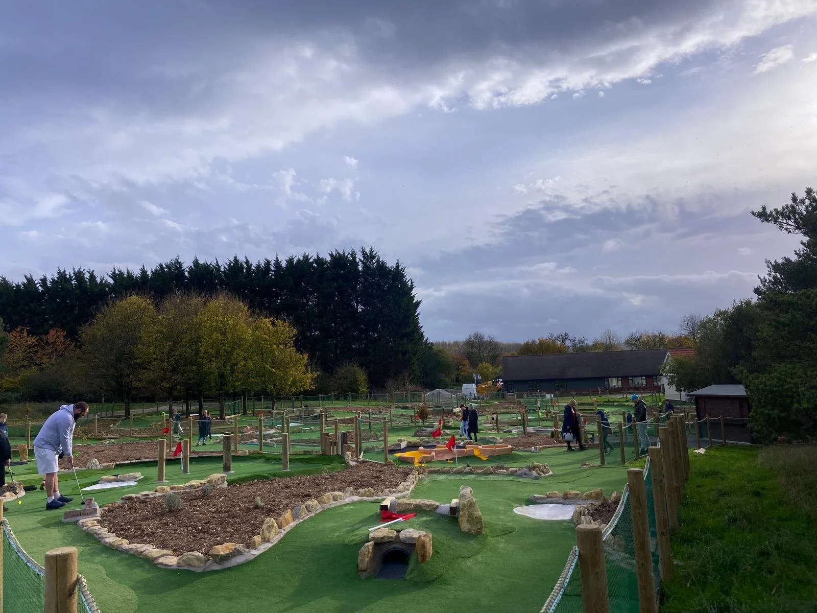 A group of people playing mini golf on a course with various obstacles, situated outdoors near trees and a building under a cloudy sky.