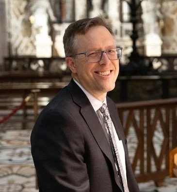 A man in a dark suit, light shirt, and patterned tie, smiling in an indoor setting with ornate decor and wooden railings.