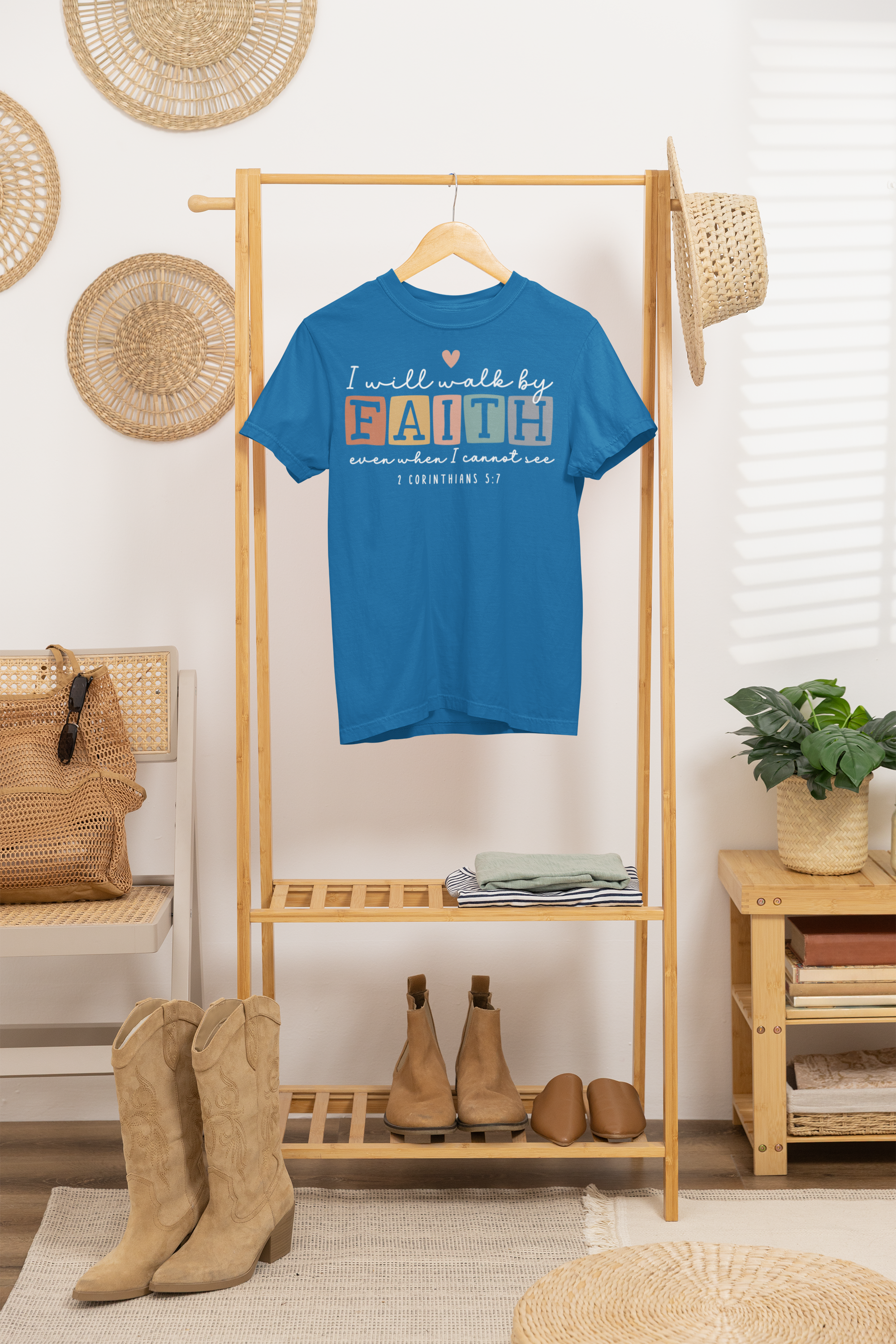 A blue T-shirt hanging on a wooden clothing rack with a religious quote about faith, surrounded by woven hats, a plant, and footwear in a cozy room.