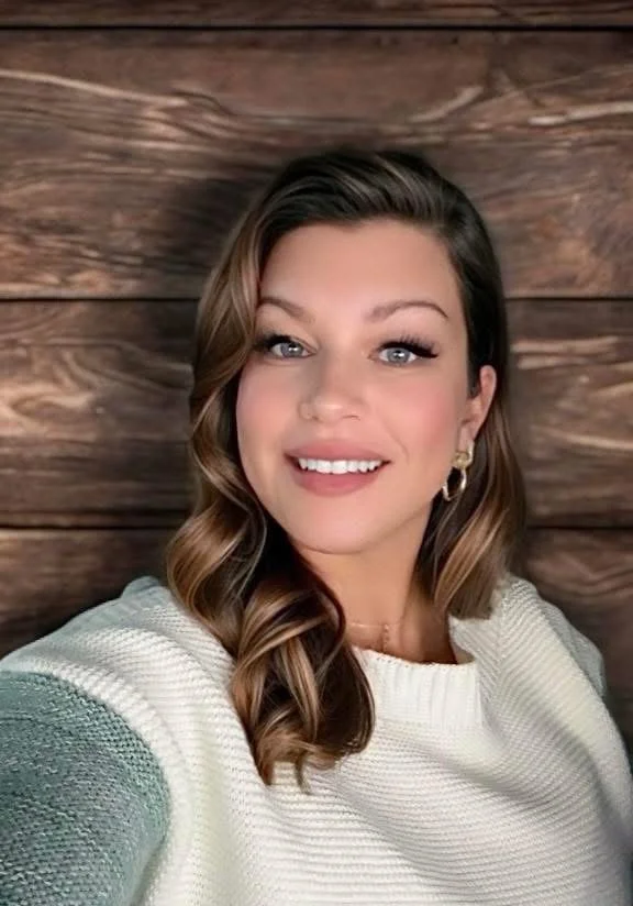 A young woman with long, wavy brown hair and blue eyes smiling, wearing a white sweater and gold earrings, against a wooden wall background.
