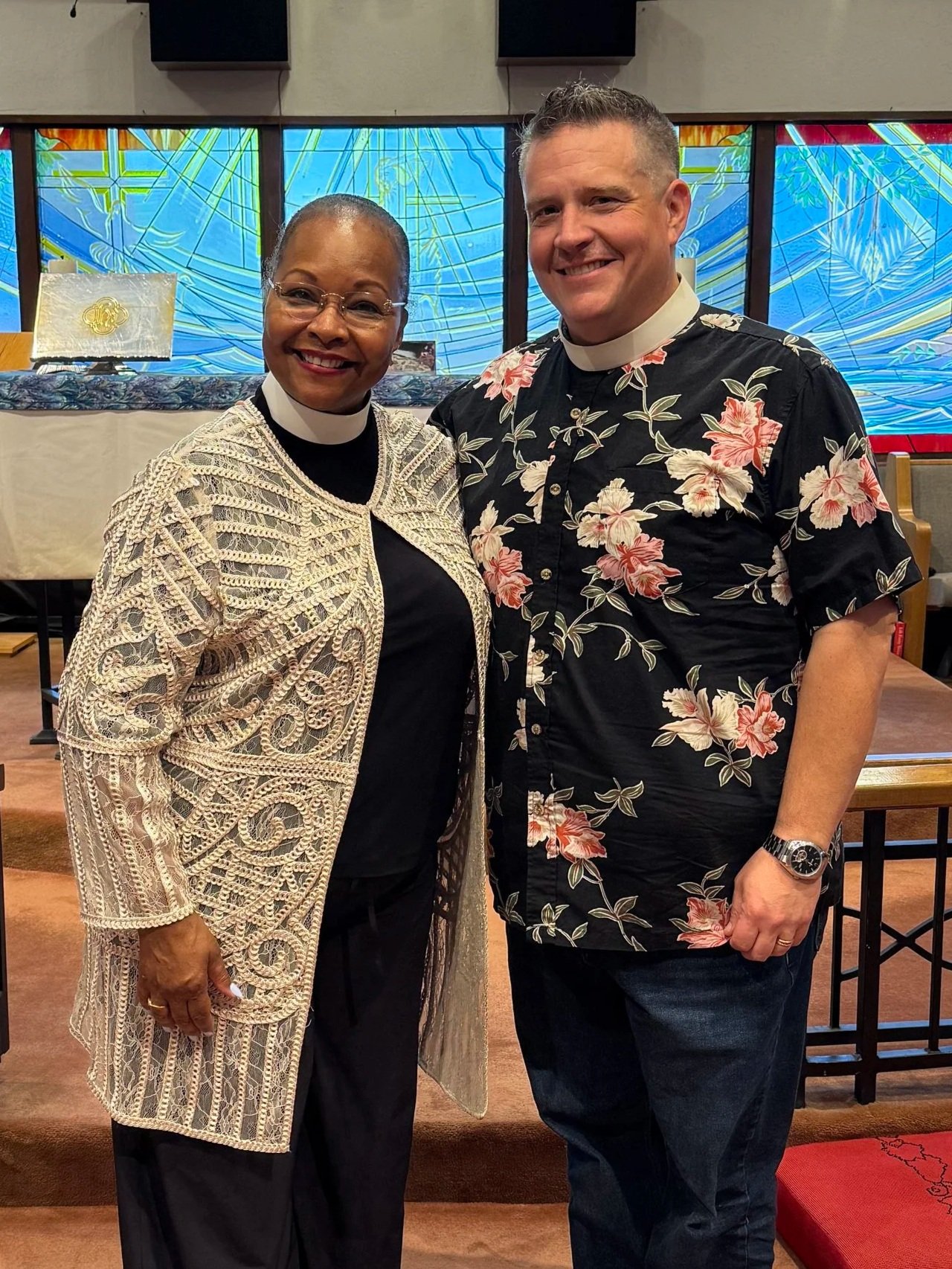 A woman and a man standing inside a church, smiling at the camera. The woman is wearing glasses, a black top, and an intricate beige crochet jacket. The man is wearing a black Hawaiian shirt with pink and white floral print and a clerical collar, indicating he is a priest. The background shows colorful stained glass windows and church furnishings.
