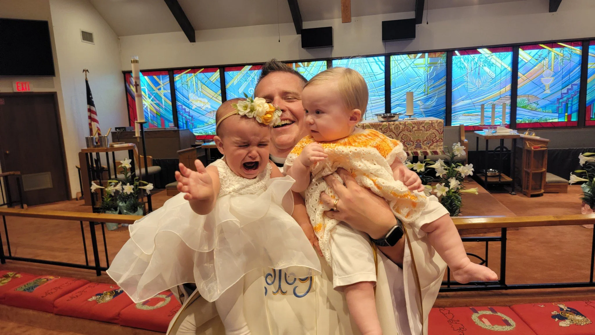 A clergy member holding two young children during a religious ceremony inside a church, with stained glass windows and floral decorations in the background.