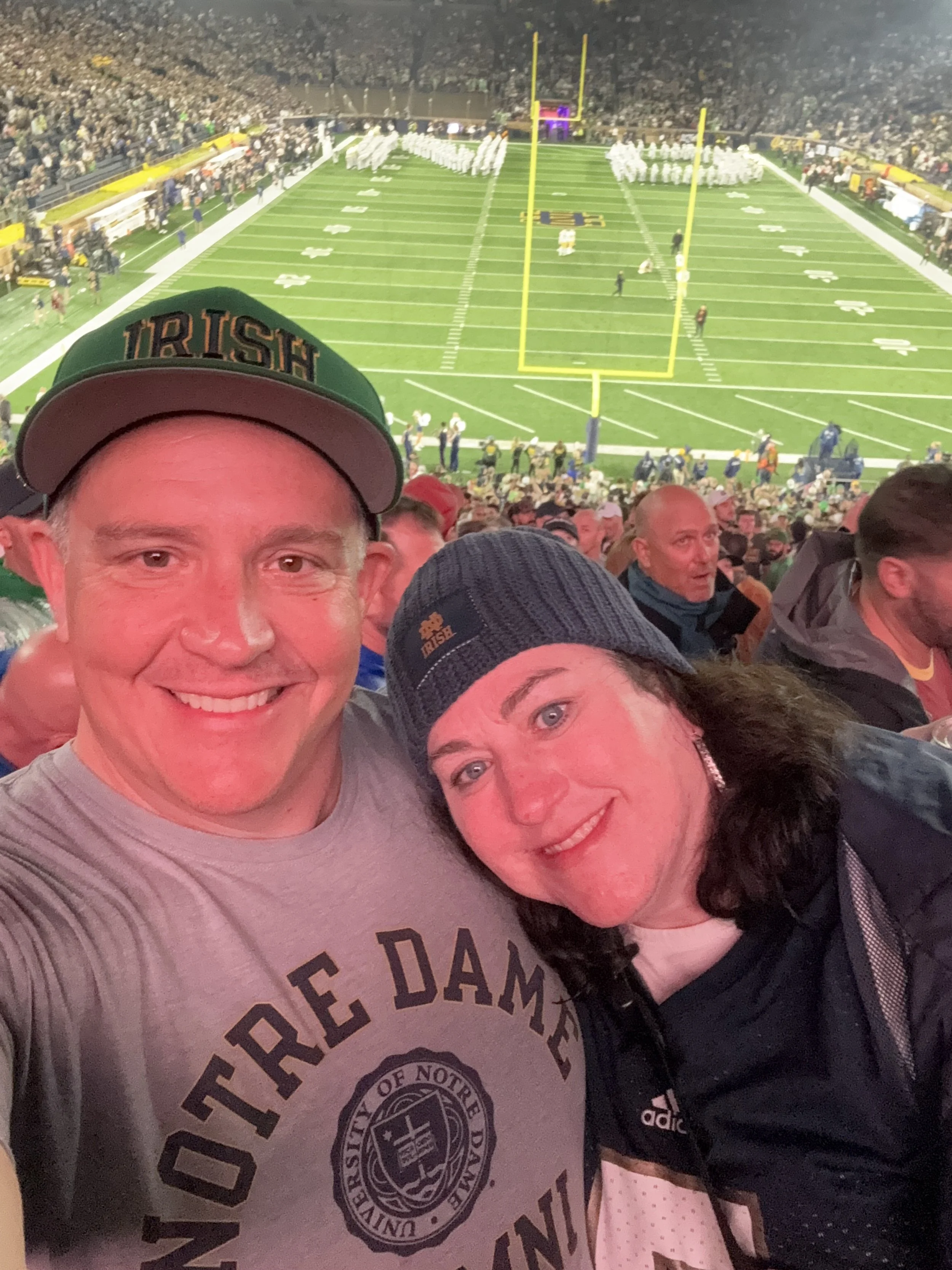 Two smiling people at a football game, taking a selfie in the stands with the field visible behind them. The man is wearing a Notre Dame shirt and a green cap, and the woman is wearing a beanie and a sports jersey.