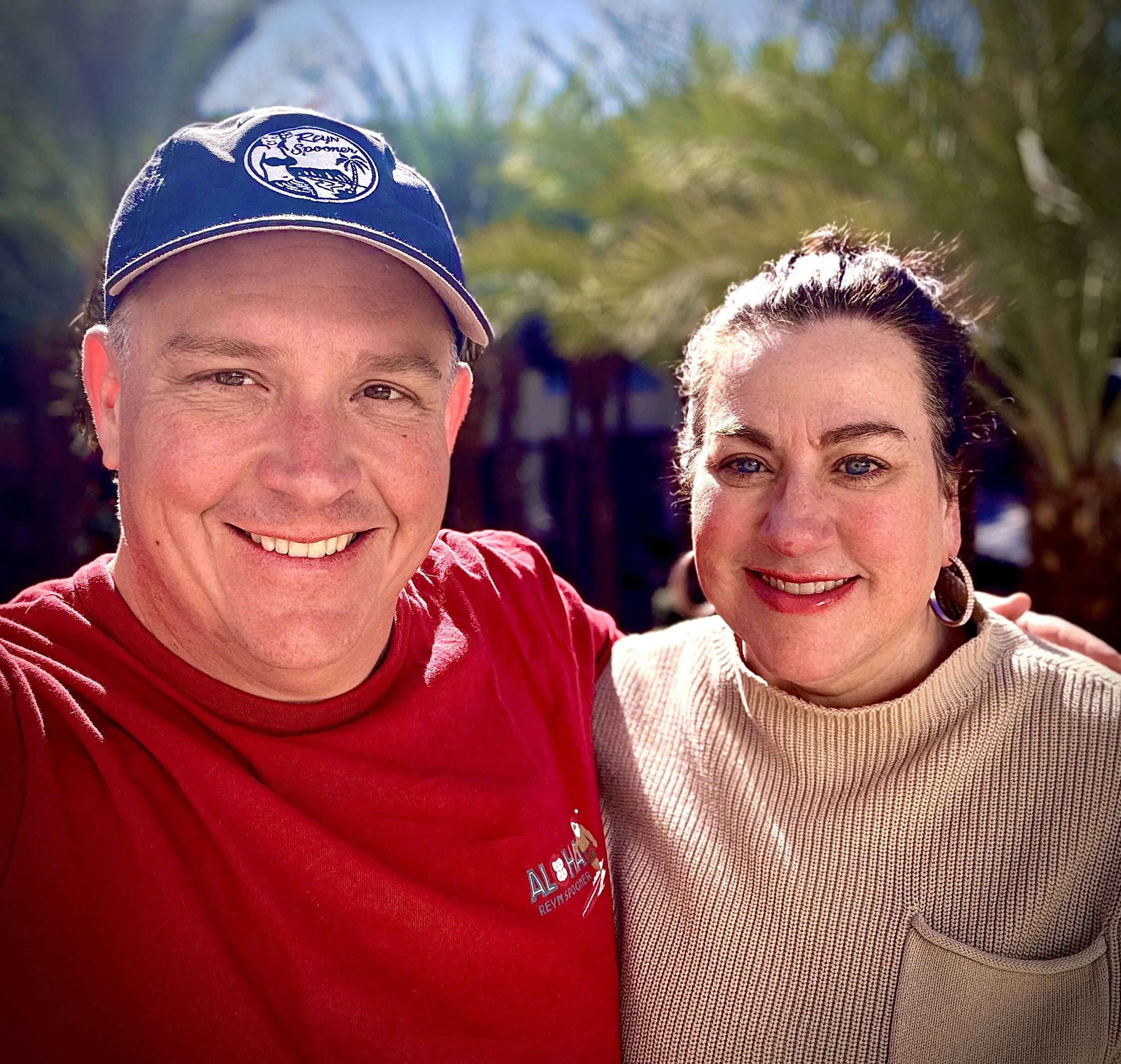 A smiling man in a blue cap and red shirt with a woman in a beige sweater, outdoors on a sunny day with green palm trees in the background.