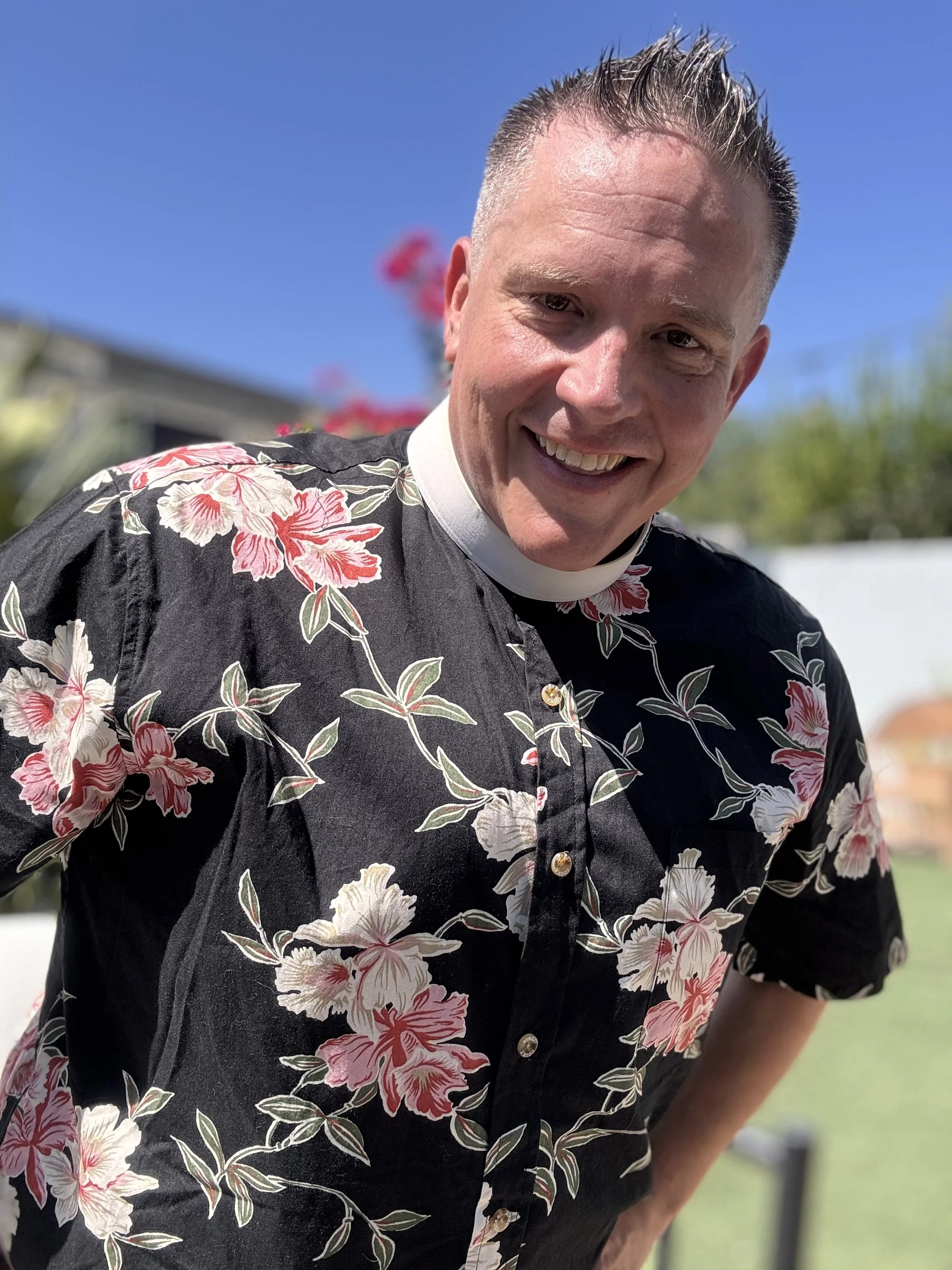 A man with short, spiked hair smiling at the camera, wearing a black shirt with pink and white floral patterns, outdoors with a background of a house, green bushes, and a clear blue sky.