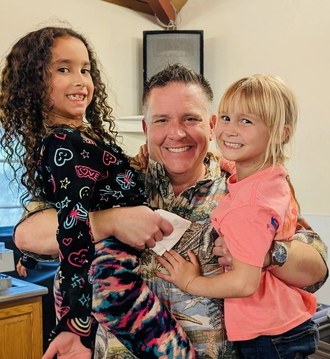 A man smiling and holding two young girls, one with dark curly hair and the other with blonde straight hair, in an indoor setting.