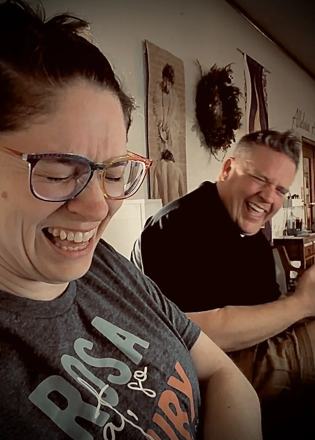 Two people laughing and smiling indoors, with artwork and wall decorations visible in the background.