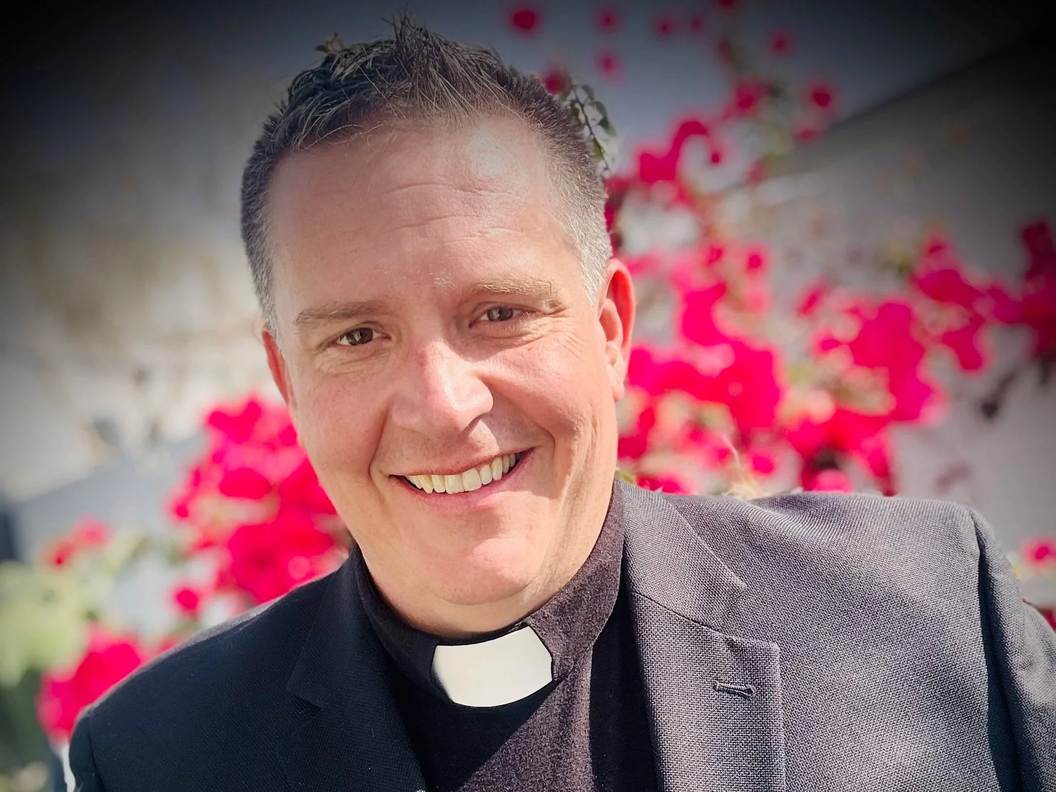 Smiling man in clerical attire with a black suit and white clerical collar, standing outdoors with pink flowers in the background.