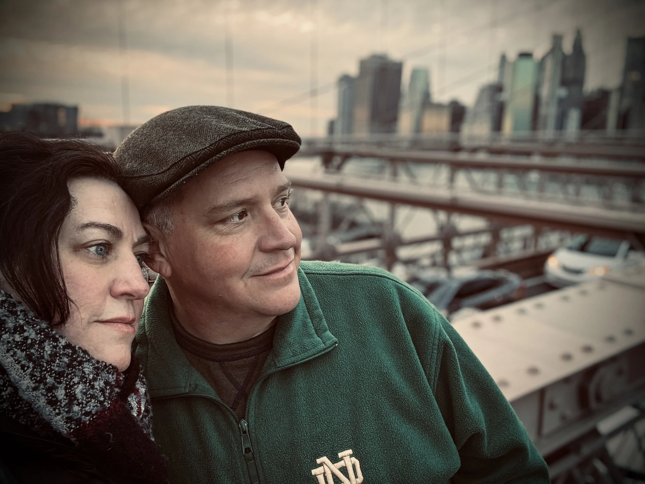 A man and a woman are outdoors, standing close together, with a city skyline and bridges in the background during sunset.