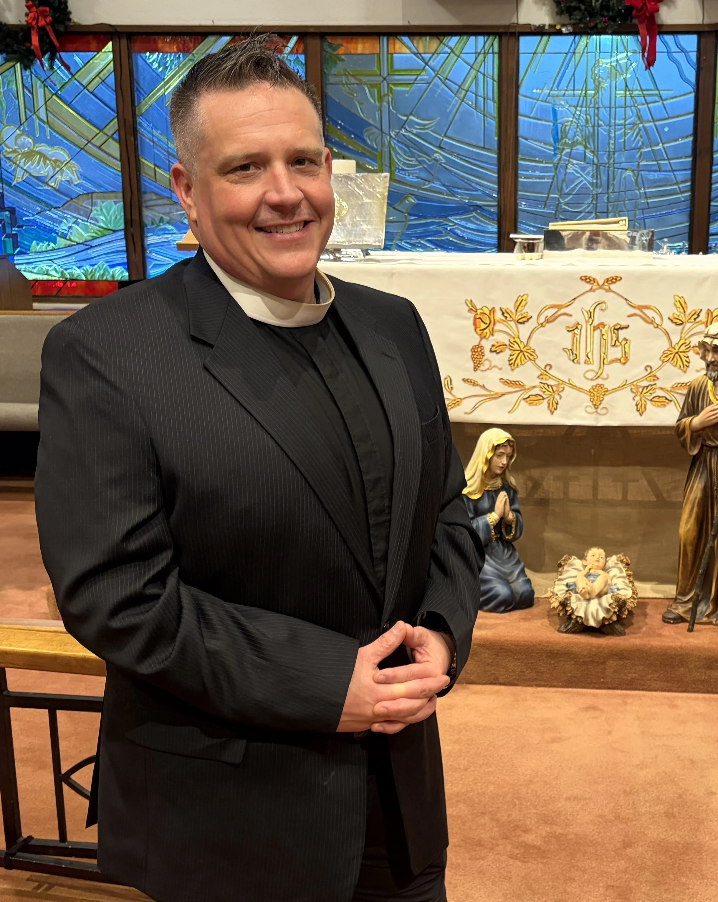 A smiling man in a black clerical suit standing in a church with religious statues and stained glass windows in the background.