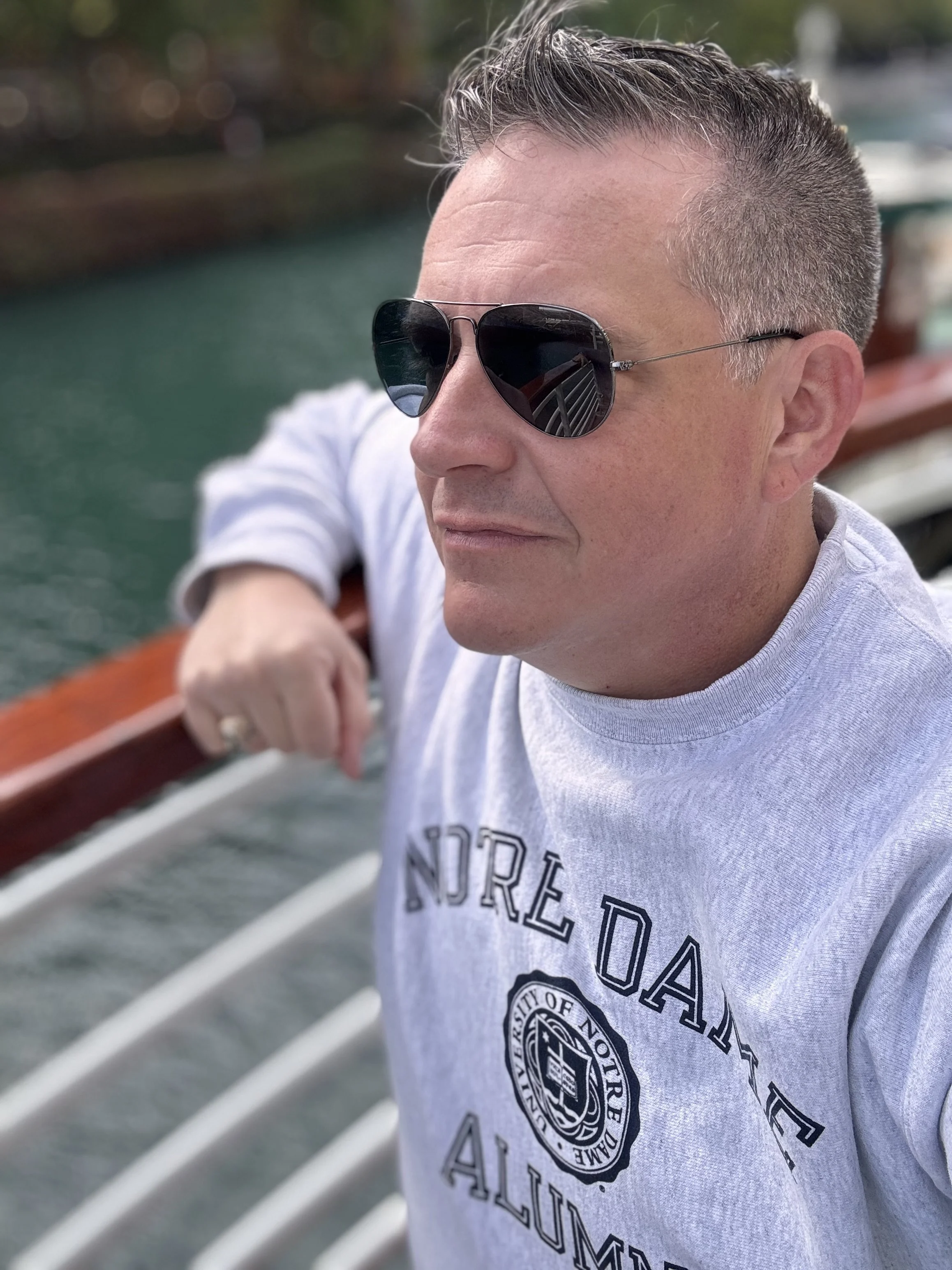 A man wearing sunglasses and a gray sweatshirt with 'Northgate Alabama' printed on it, leaning on a boat railing with water in the background.