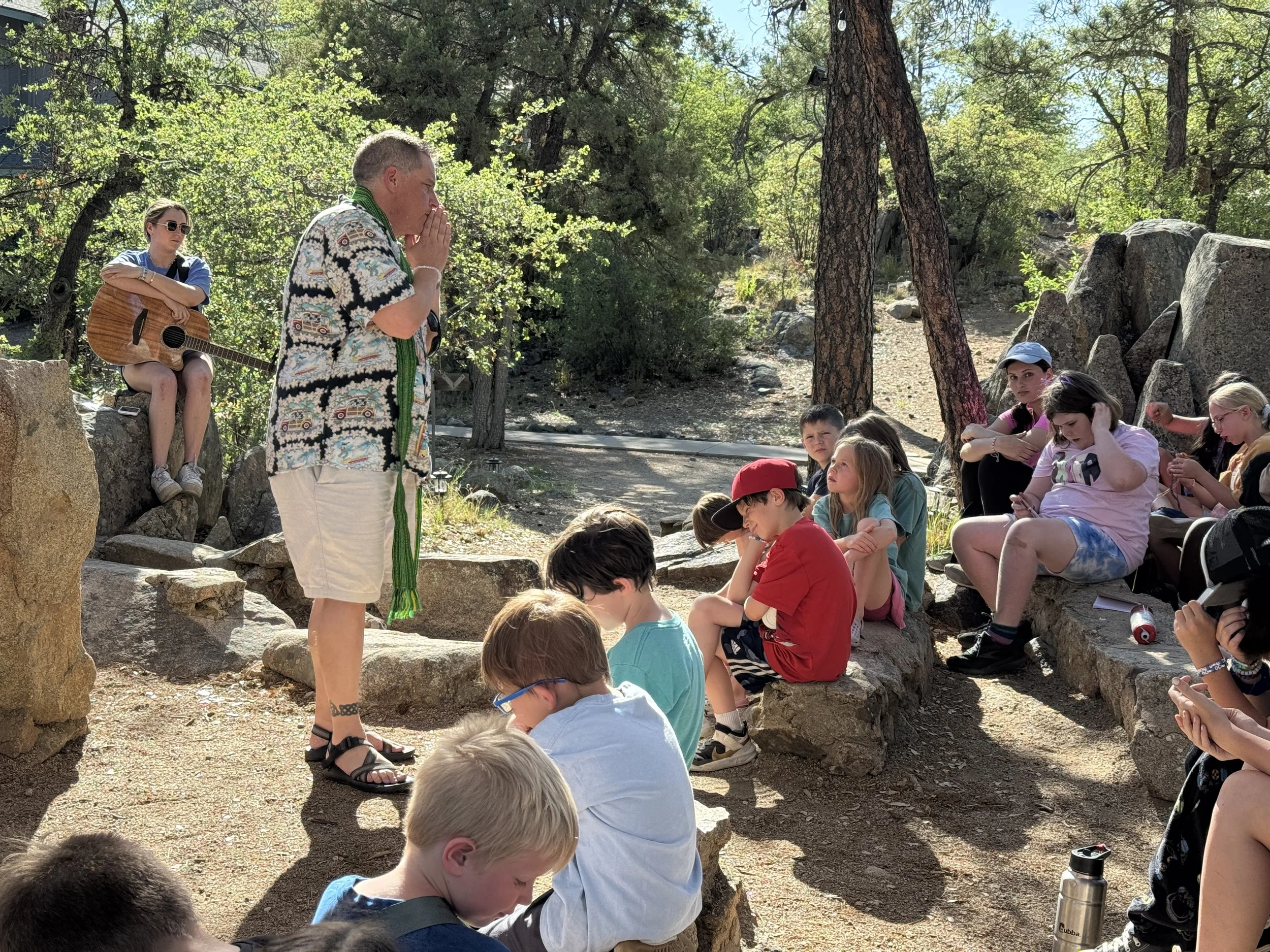 Man standing and praying with children seated on rocks around him outdoors, some children are using phones or playing, with trees and rocks in the background.