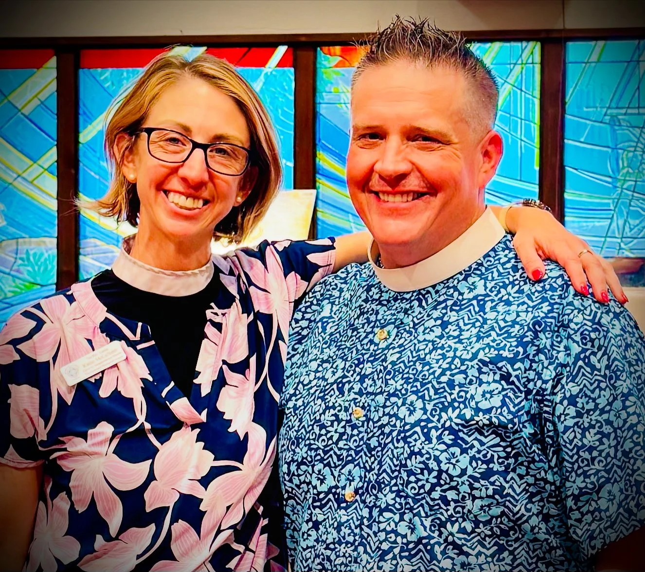 A woman and a man smiling at the camera, with the woman wearing glasses and a name badge, and the man wearing a blue patterned shirt, in front of colorful stained glass windows.