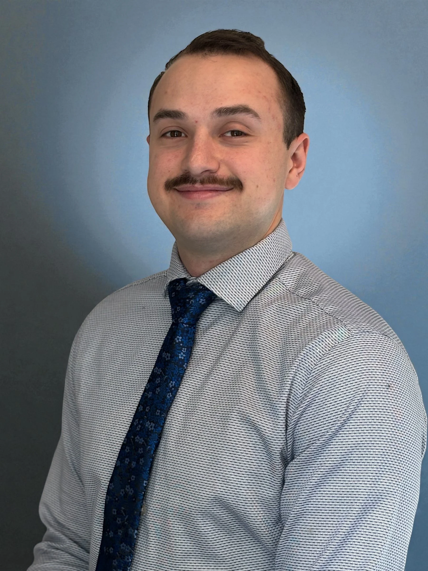 A man with a mustache and short dark hair smiling at the camera, wearing a light patterned dress shirt and a dark blue tie with floral pattern, standing against a blue wall.