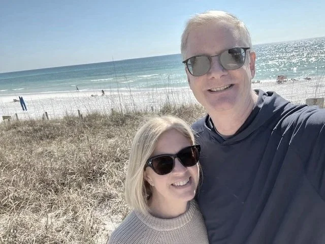 A man and woman wearing sunglasses taking a selfie on a beach with sand, some grass, and the ocean in the background.