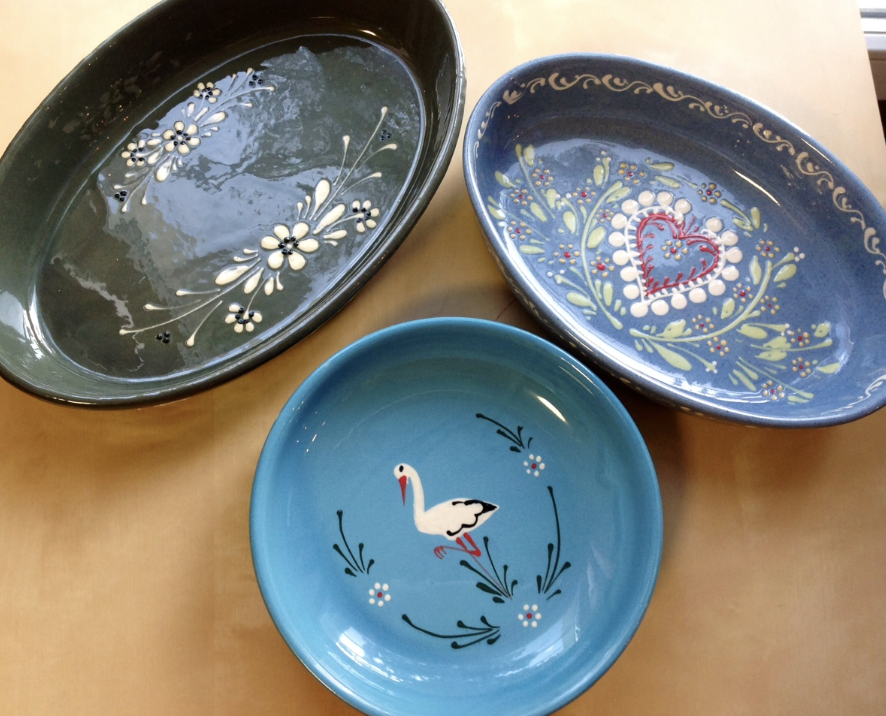 Three decorative ceramic bowls on a wooden table, each with floral and nature-themed patterns. One bowl contains water, and they are arranged with one blue bowl featuring a white stork design, a darker blue bowl with a red heart and floral pattern, and a dark green bowl with white floral motifs.