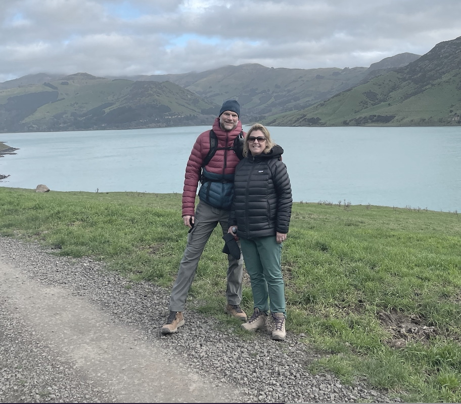 Two people stand together on a gravel path near a lake, with green hills and mountains in the background, dressed in outdoor hiking gear.