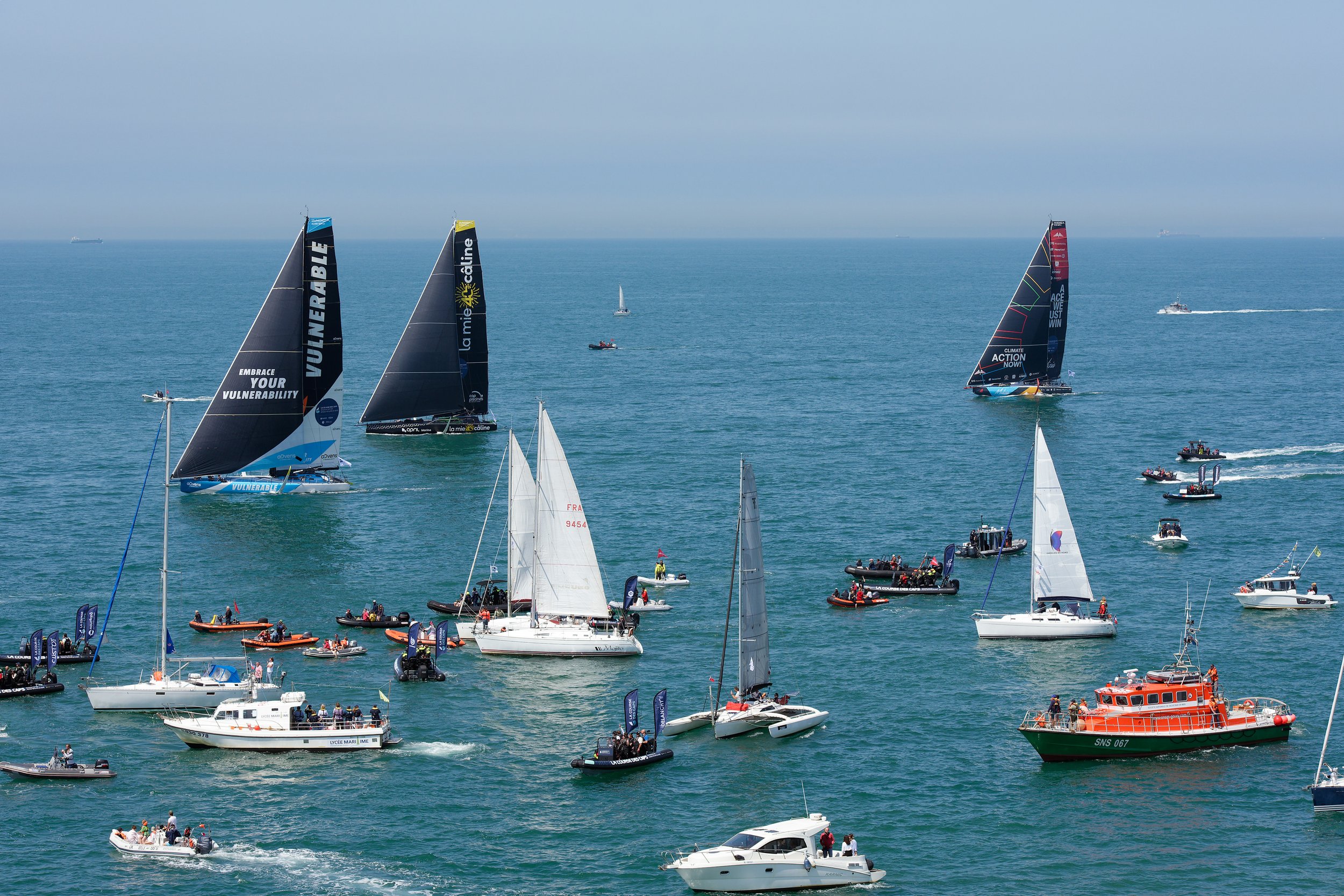 Plusieurs voiliers et bateaux de différentes tailles en mer, certains avec des voiles déployées, dans une journée ensoleillée.
