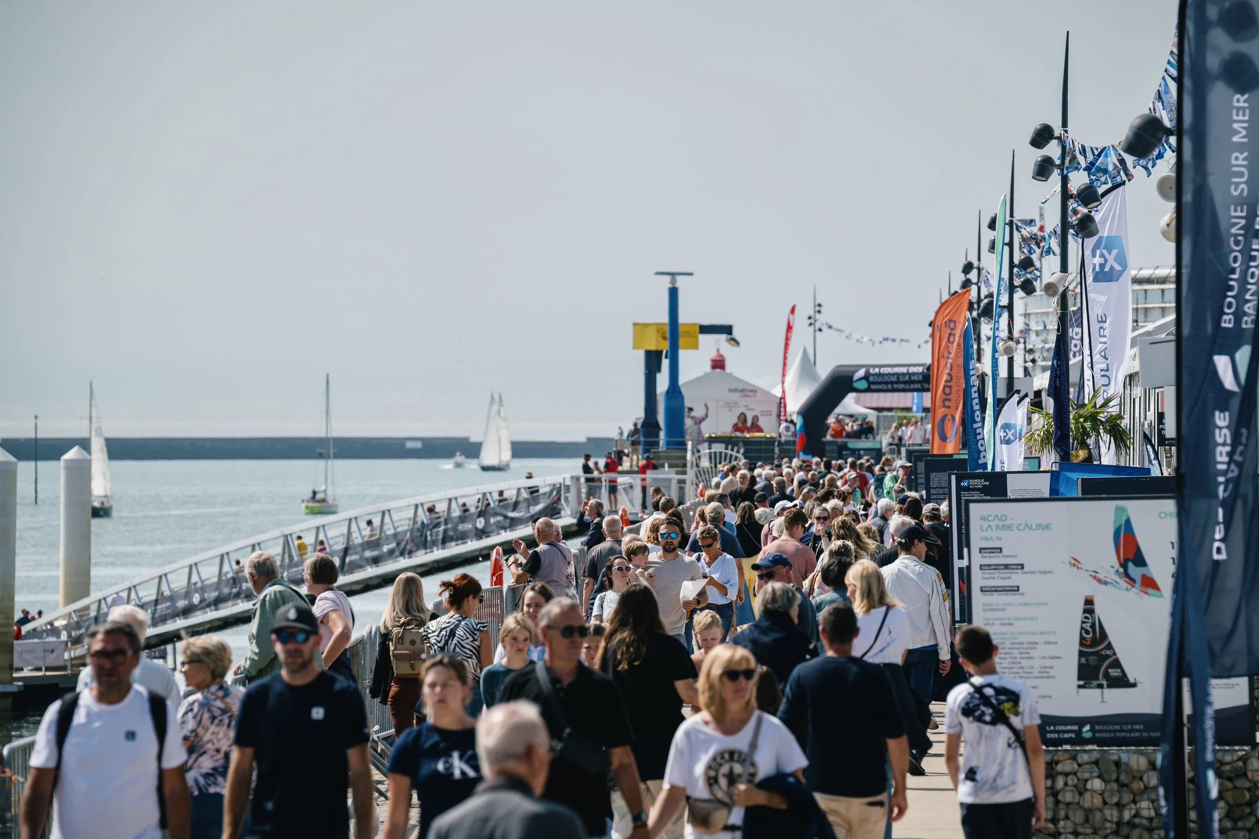 Une foule de personnes marche sur une jetée au bord de la mer, avec plusieurs voiliers en arrière-plan et des drapeaux publicitaires le long de la promenade.