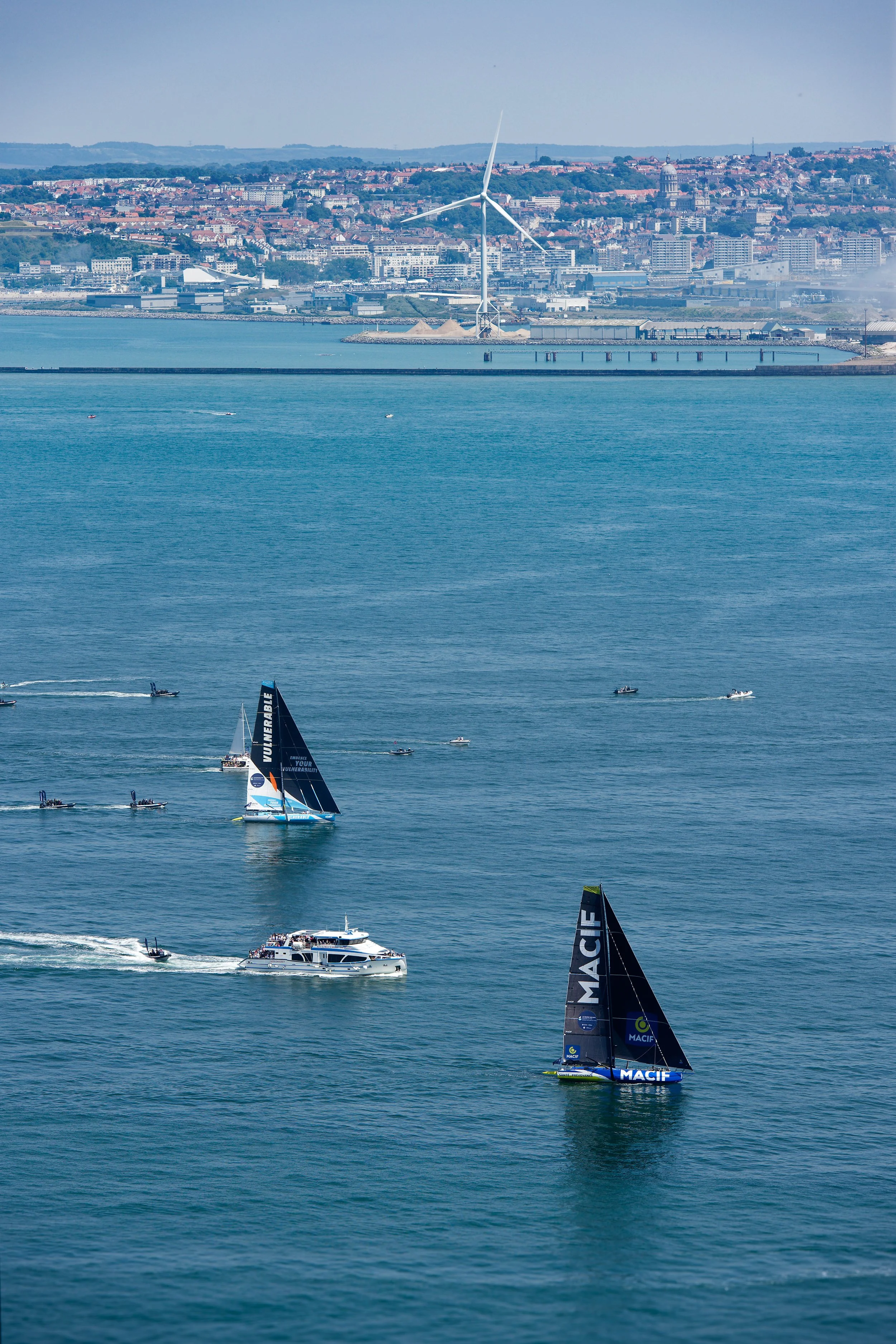 Voiliers et bateaux naviguant en mer devant une ville avec des bâtiments et un parc éolien en arrière-plan.