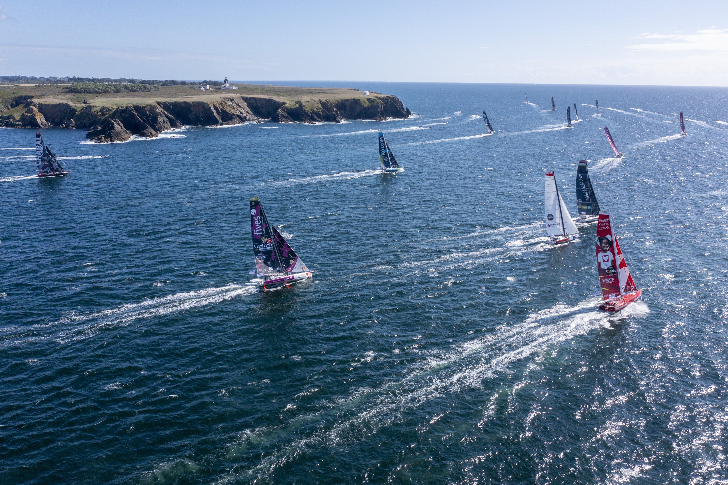 Voiliers de course naviguant en mer, avec une côte rocheuse et un phare au fond.