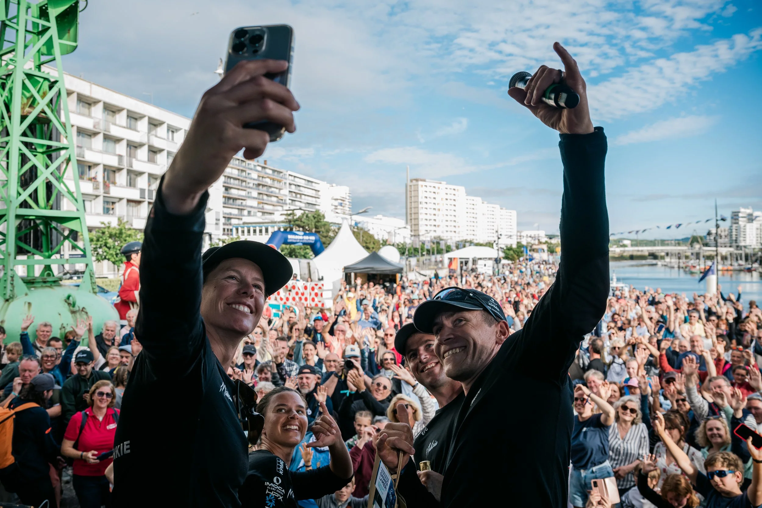 Groupe de personnes en tenue sportive prenant une selfie lors d'un événement en extérieur avec une foule nombreuse en arrière-plan et des bâtiments modernes, en journée avec ciel partiellement nuageux.