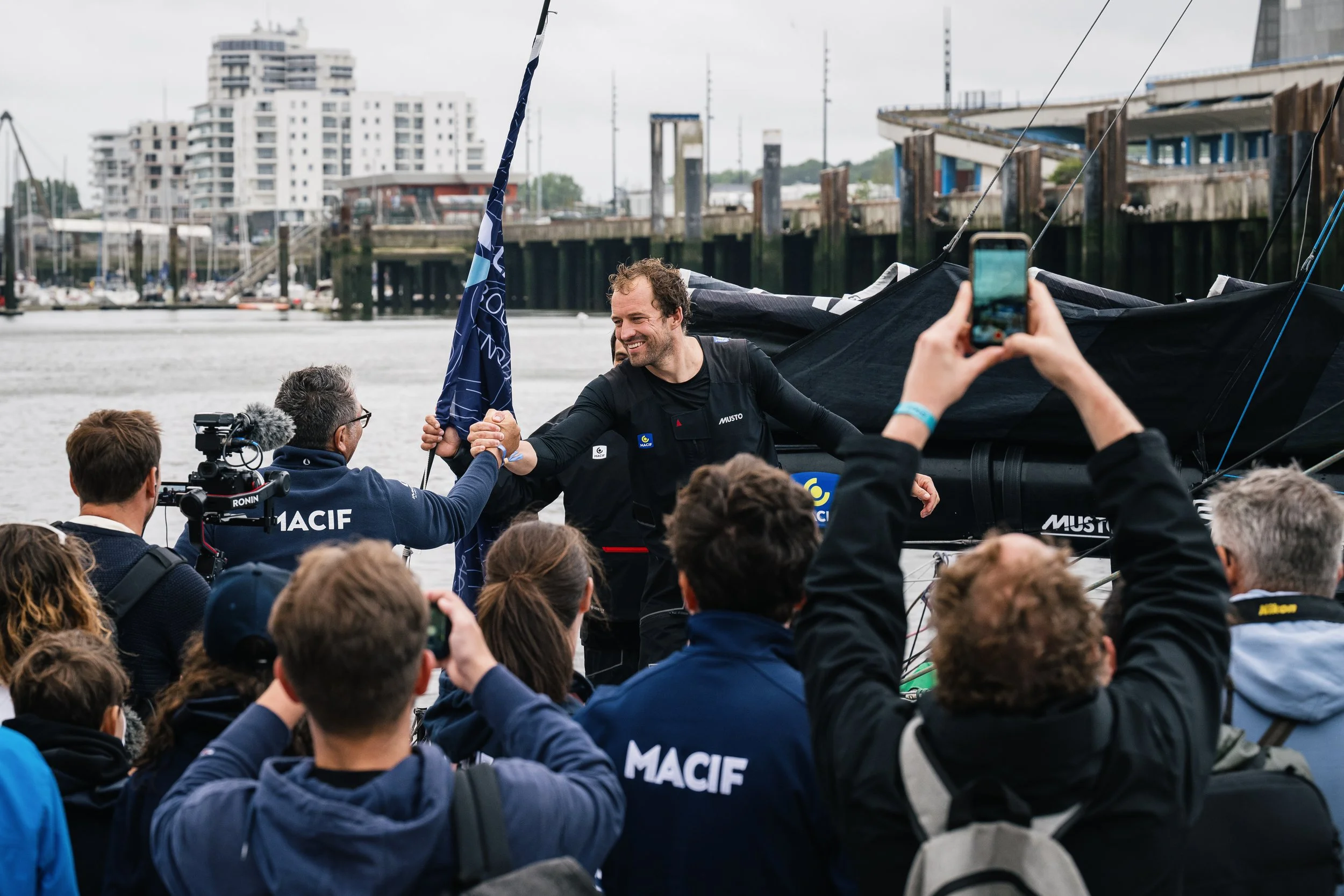 Une personne en tenue de voile serre la main d'un autre homme lors d'une cérémonie en extérieur près d'un plan d'eau, entourée d'une foule de spectateurs et de médias.