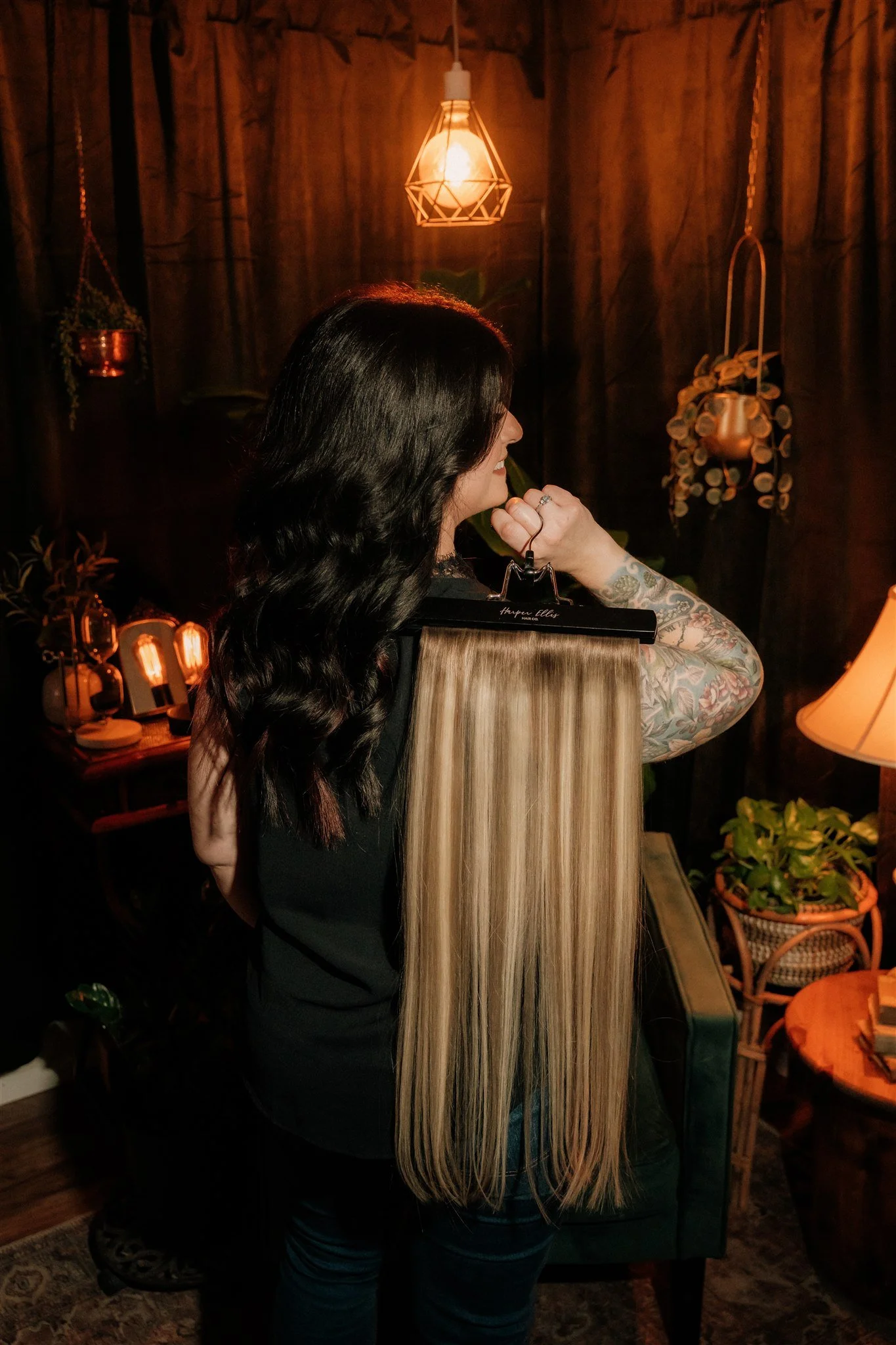 A woman with long, blonde hair and tattoos on her arm stands in a dimly lit room, holding a hair color sample swatch. The room features warm lighting, plants, and decorative items.