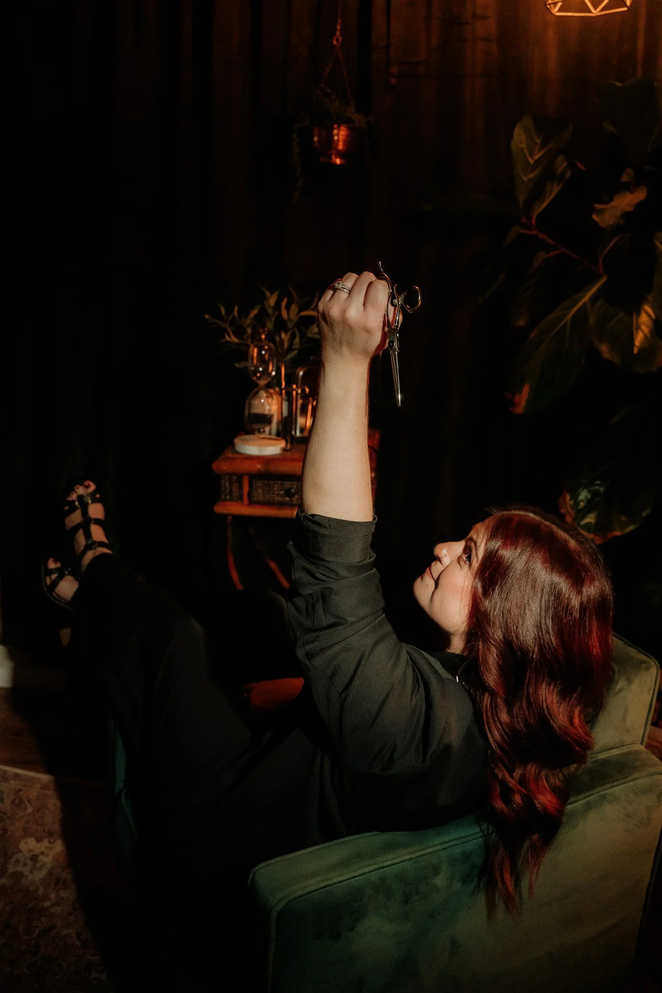 A woman with reddish hair lying in a green armchair, holding up keys in her right hand against a dark background with plants and warm lighting.