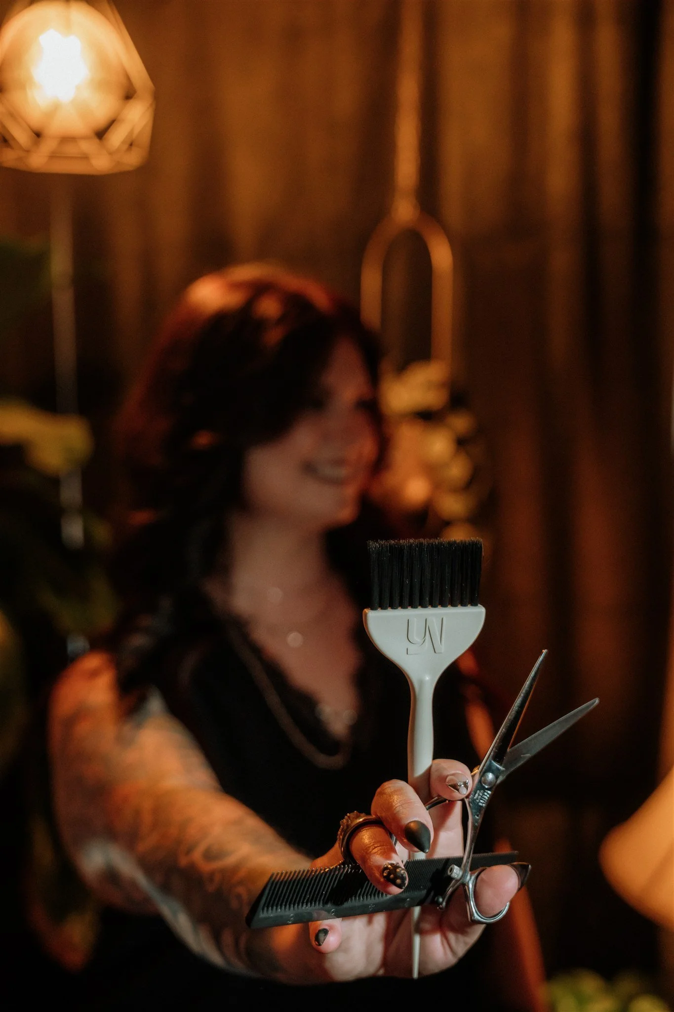 A woman with dark hair, tattoos on her arm, and black nail polish is holding hair styling tools, including a comb, scissors, and a brush, in her hand.