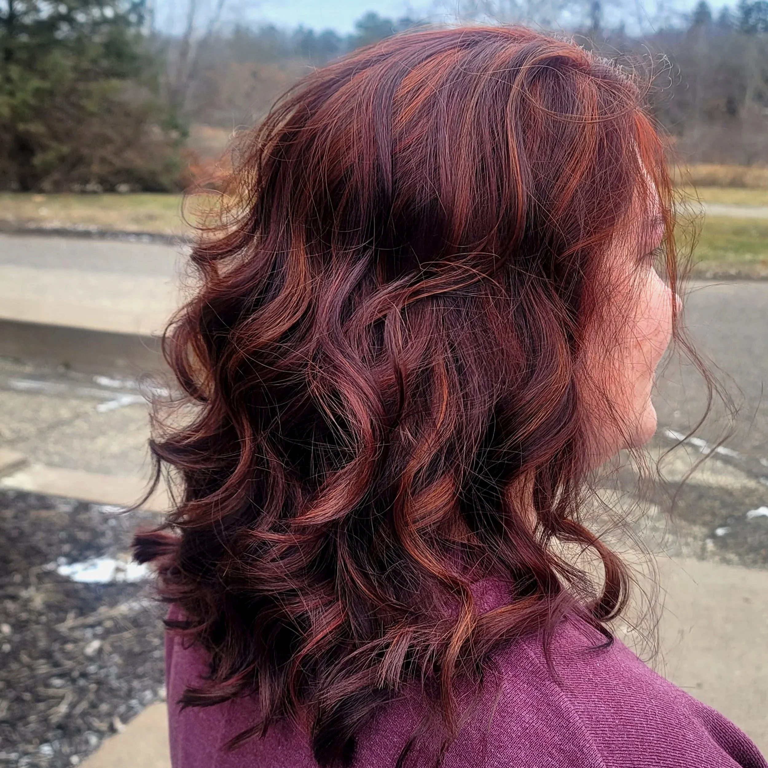 Side view of a woman with wavy, reddish-brown hair outdoors near a park or natural area.