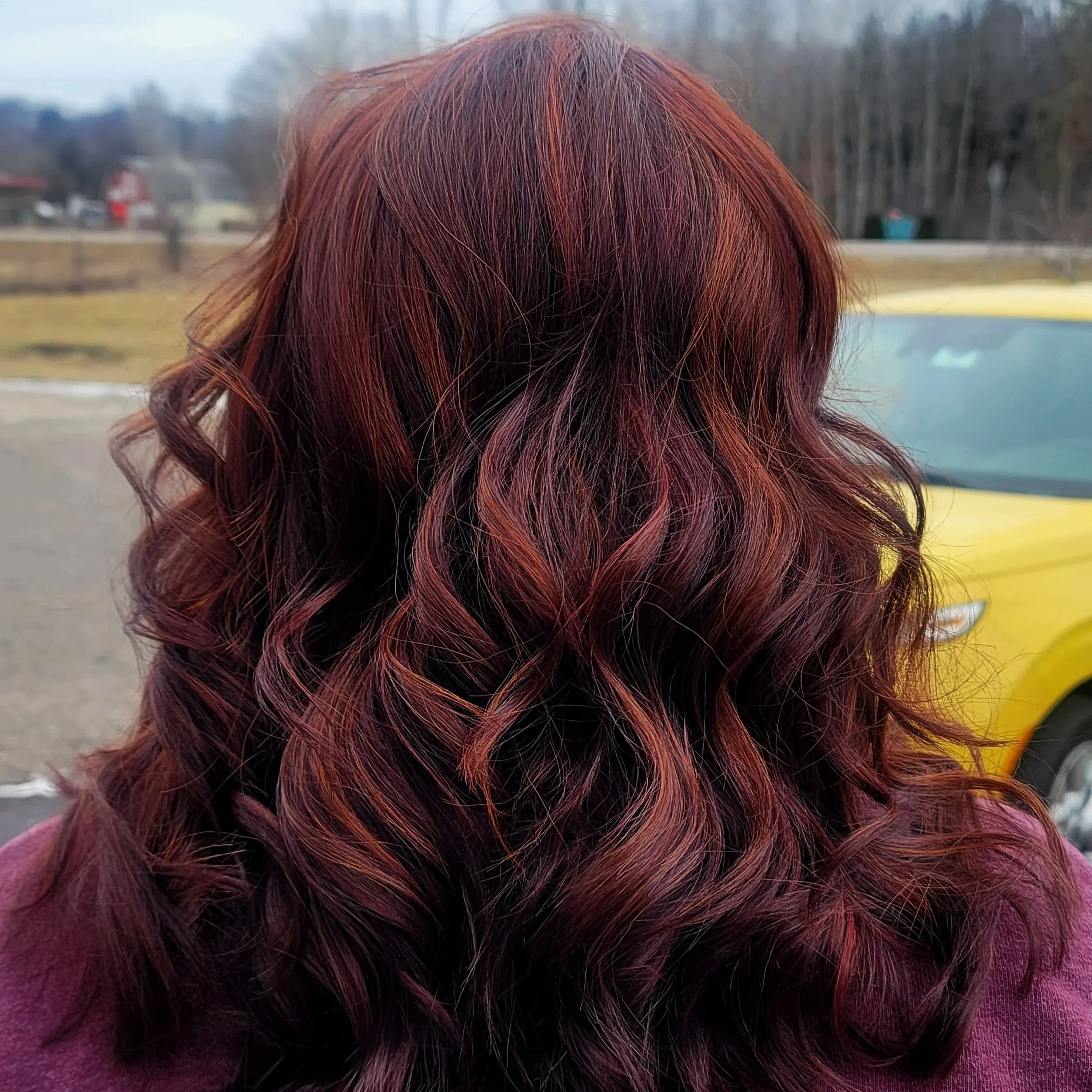 Back view of a woman with long, curly hair dyed in shades of red and purple, outdoors near a yellow vehicle.