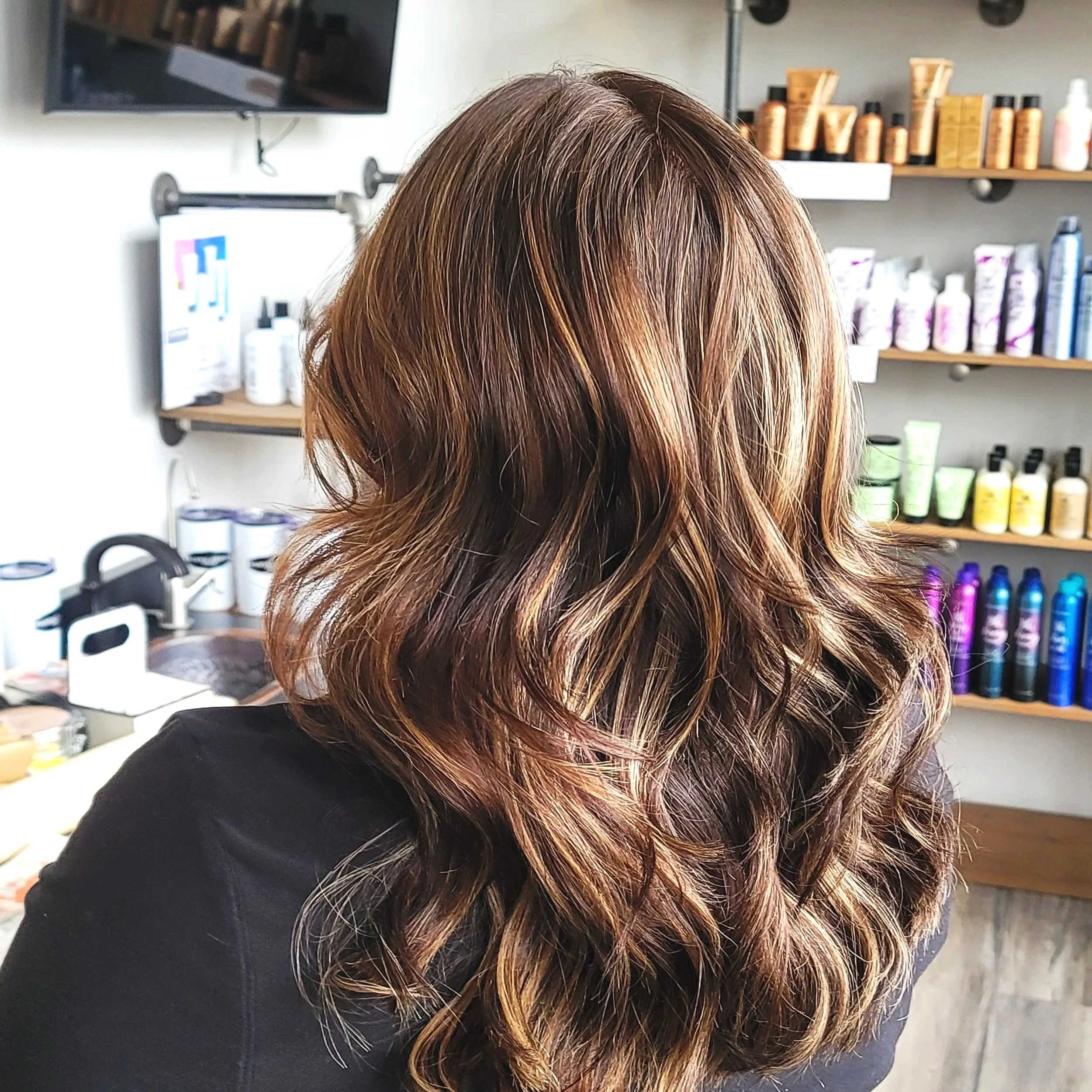 Back view of a woman's medium-length wavy brown hair with caramel highlights in a salon setting.