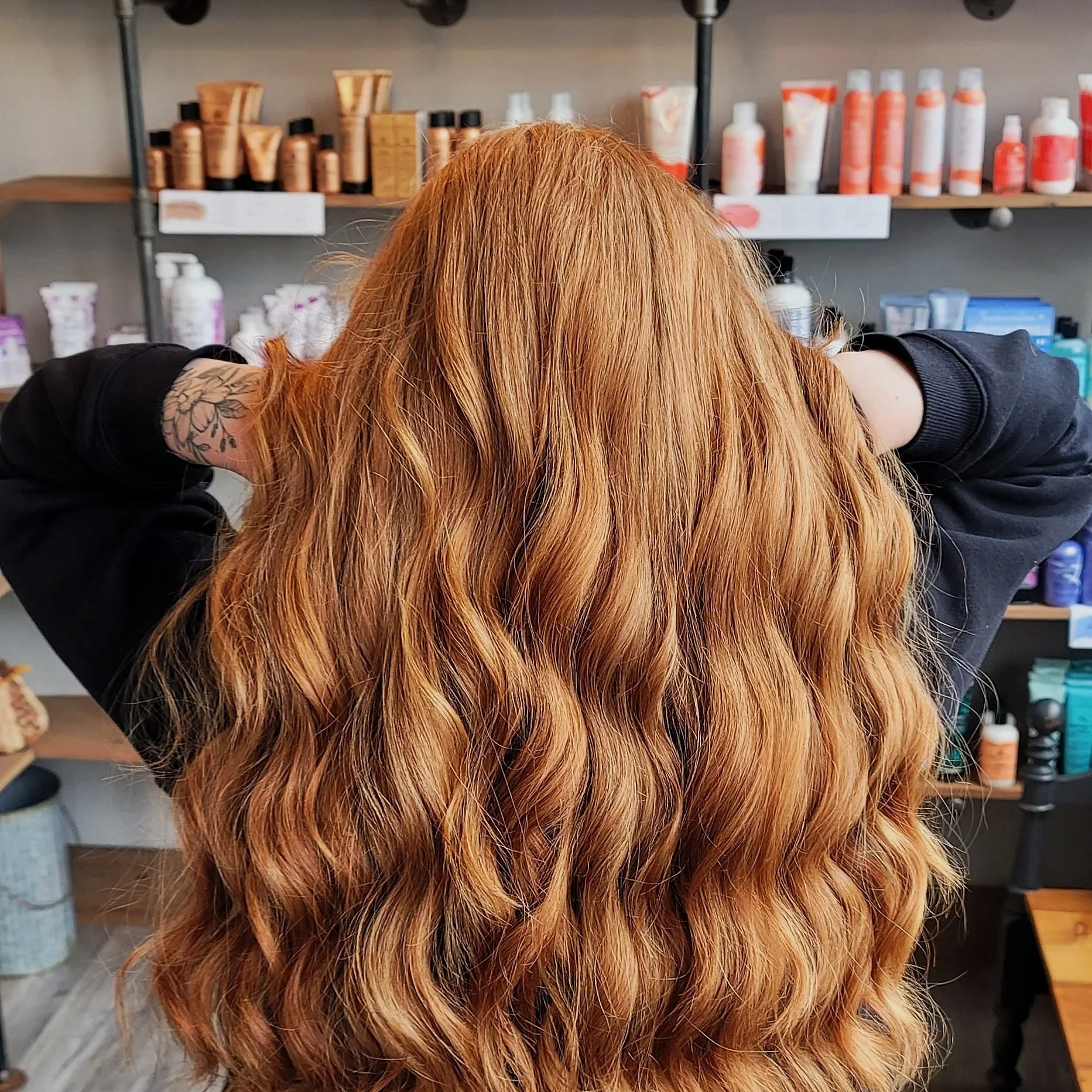 Person with long, wavy, red hair in a salon with shelves of hair products in the background.