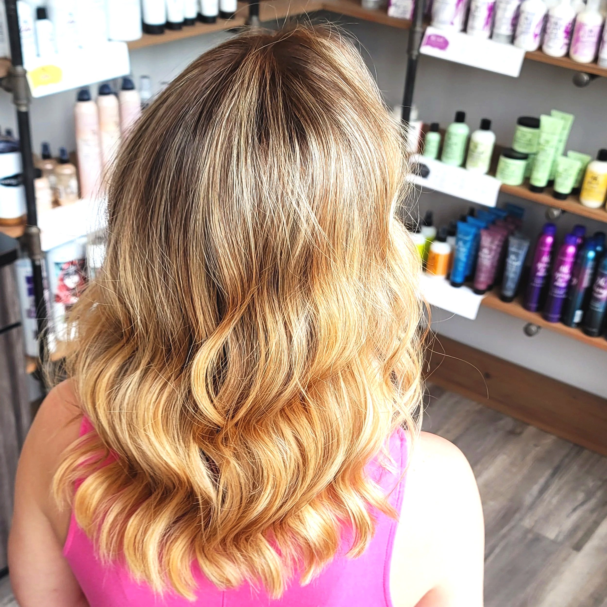 Woman with shoulder-length blonde hair styled in loose waves, wearing a pink top, in a store with shelves of hair care products.
