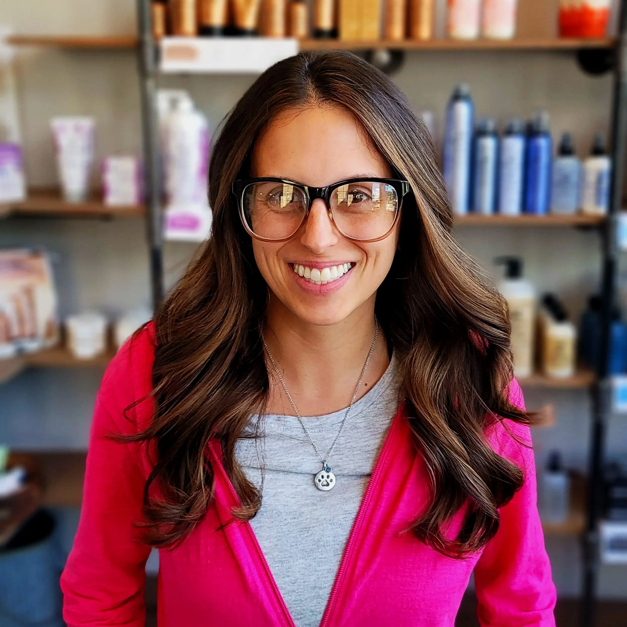 Smiling woman with long brown hair and glasses, wearing a pink jacket and a necklace with a skull pendant, standing in front of shelves with hair or skincare products.
