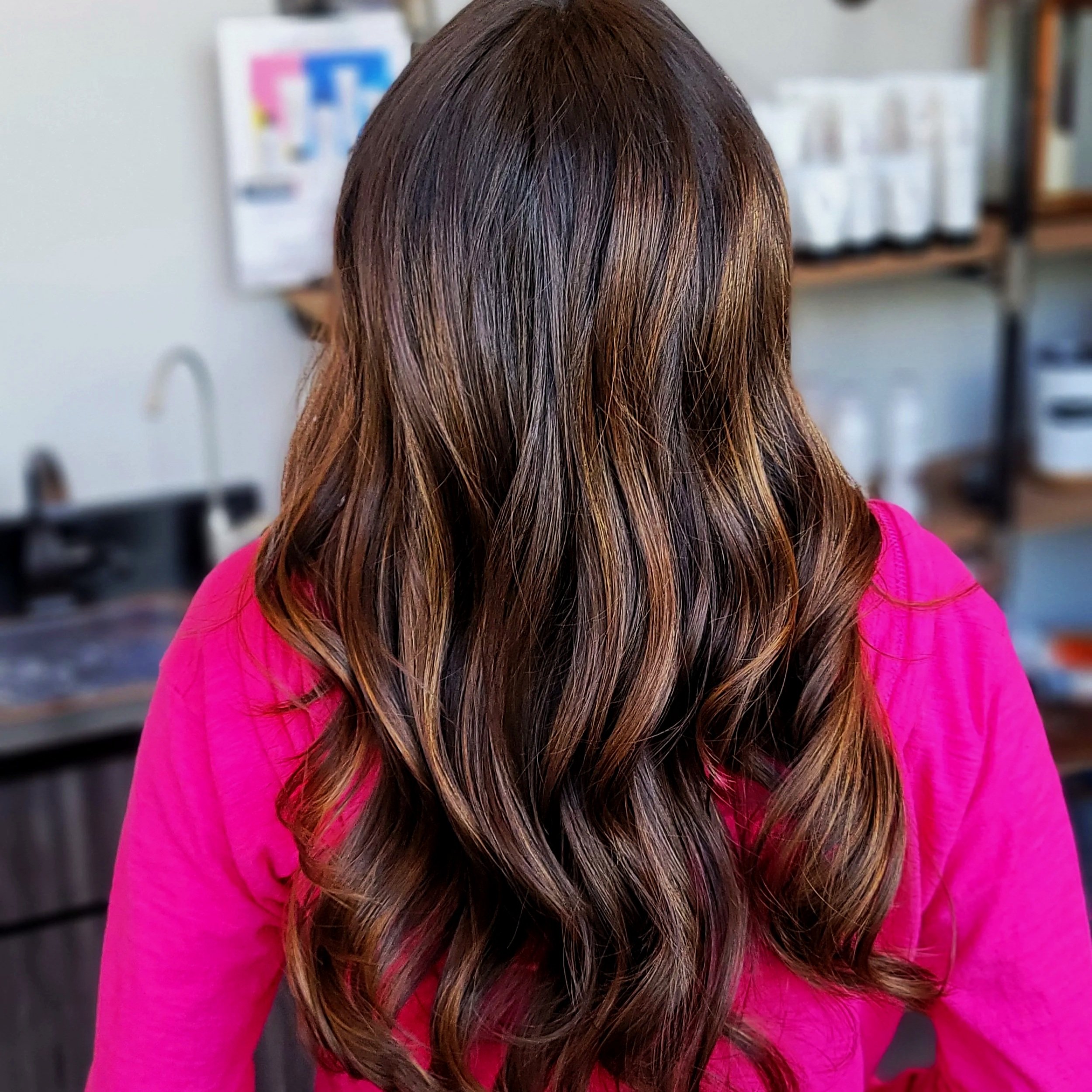 Back view of a woman with long, wavy brown hair, wearing a bright pink shirt, standing in a hair salon.