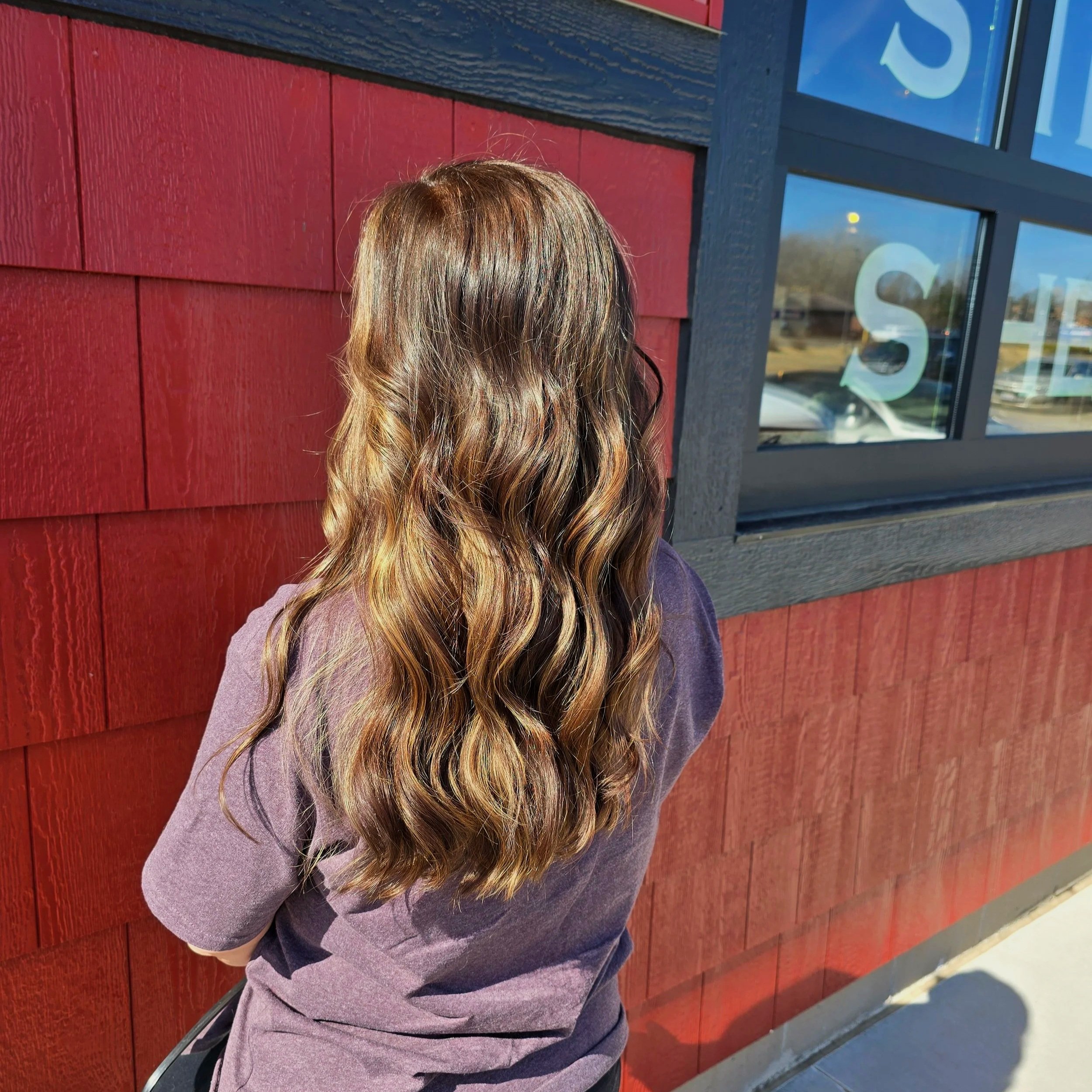 Person with long, wavy brown hair standing outside a building with red shingle siding and a window that reads 'SALE.'