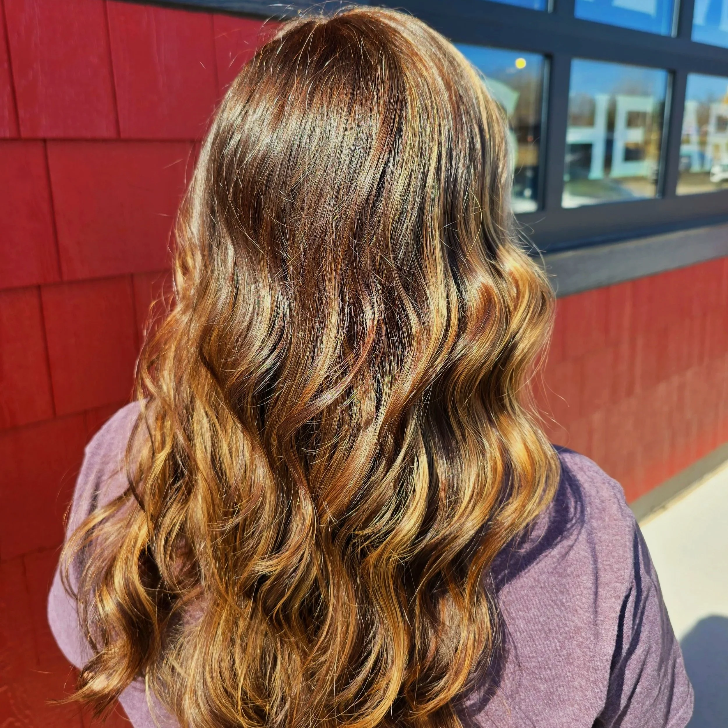 Back of a person's head with long wavy hair with shades of brown and blonde, sitting outdoors near a red wooden wall and a window.