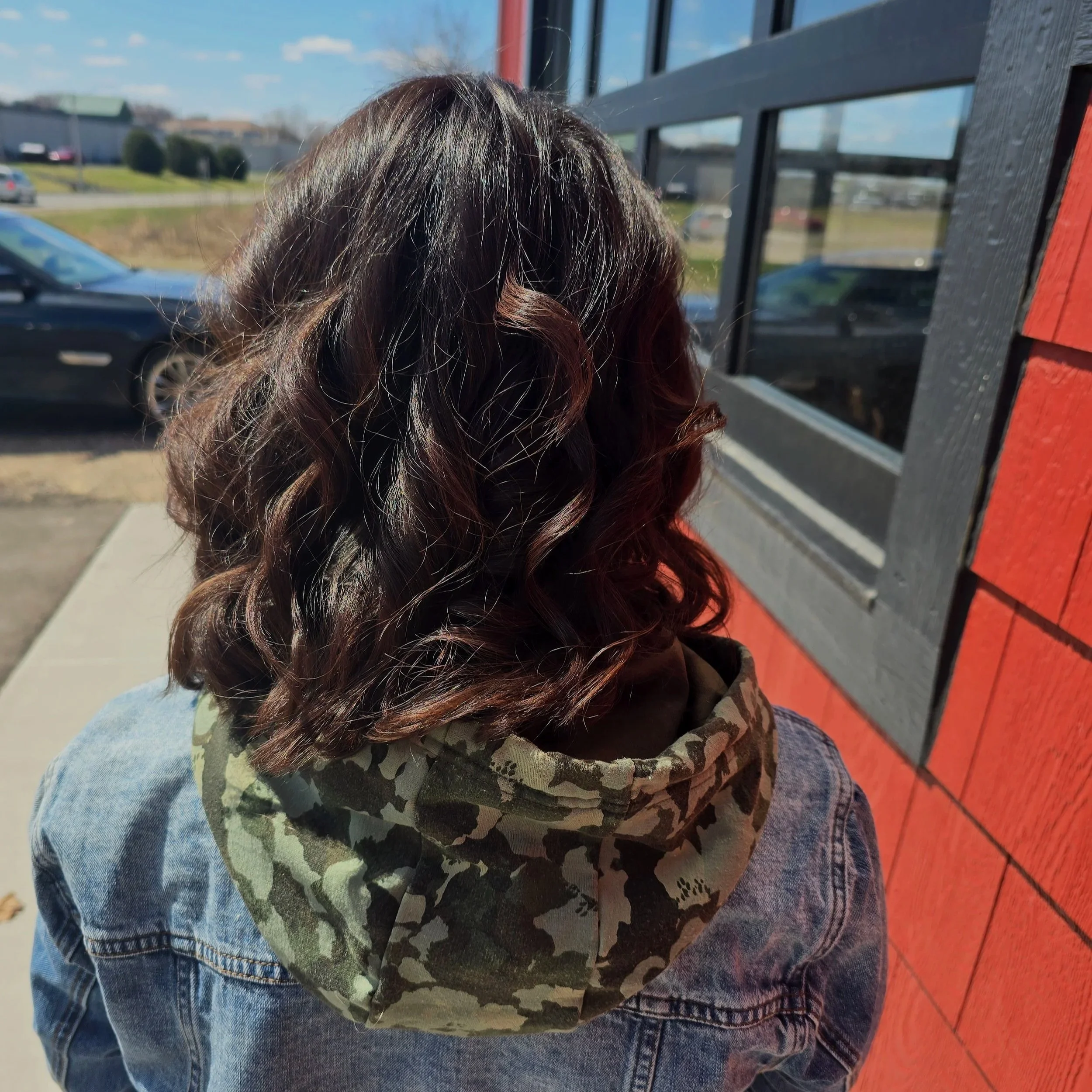 Back of a woman with curly, shoulder-length hair wearing a camouflage hoodie and denim jacket, standing outside near a red building with black window frames.