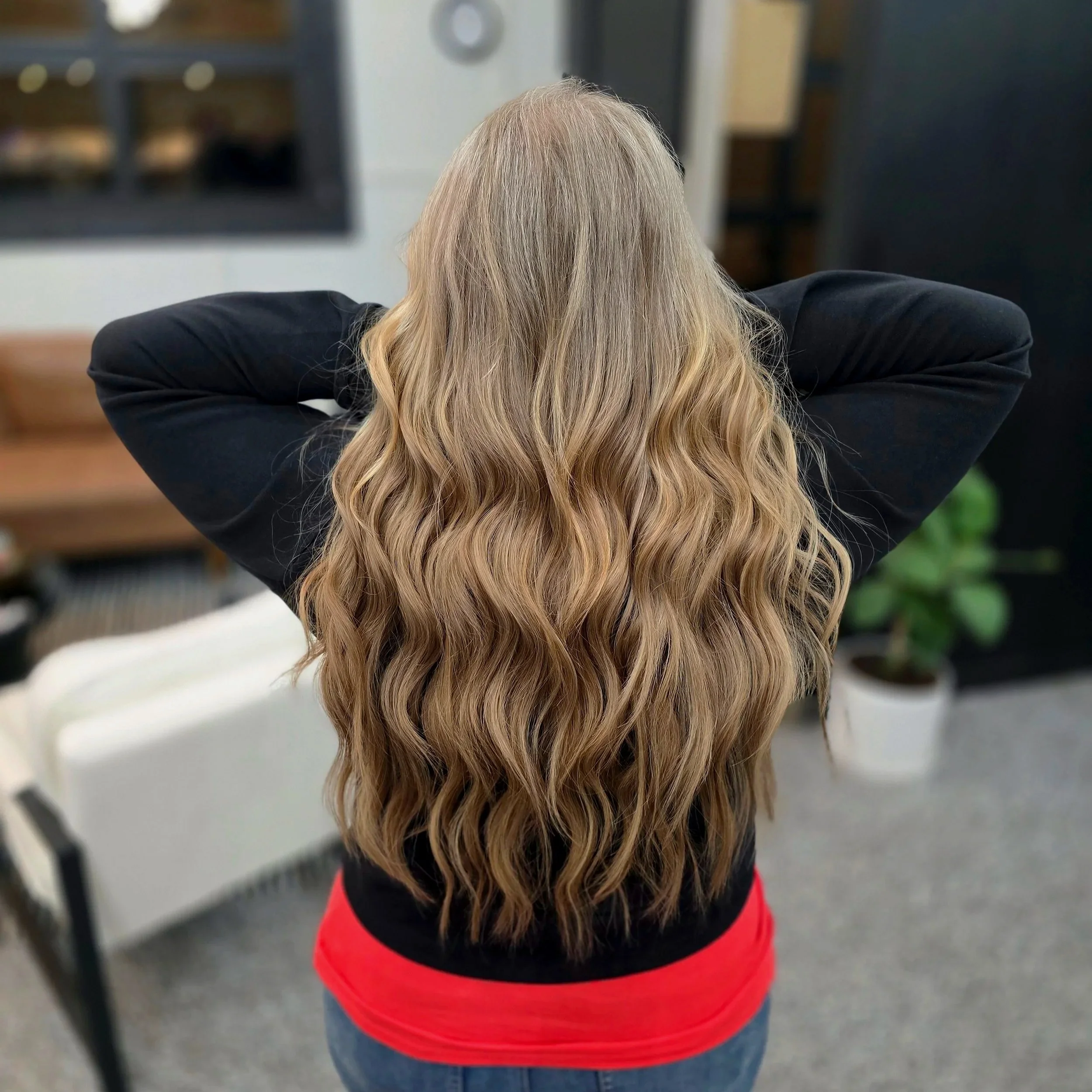 Back view of a woman with long, wavy, blonde hair wearing a black top and red shirt underneath, with her arms behind her head, in a room with a plant and furniture.